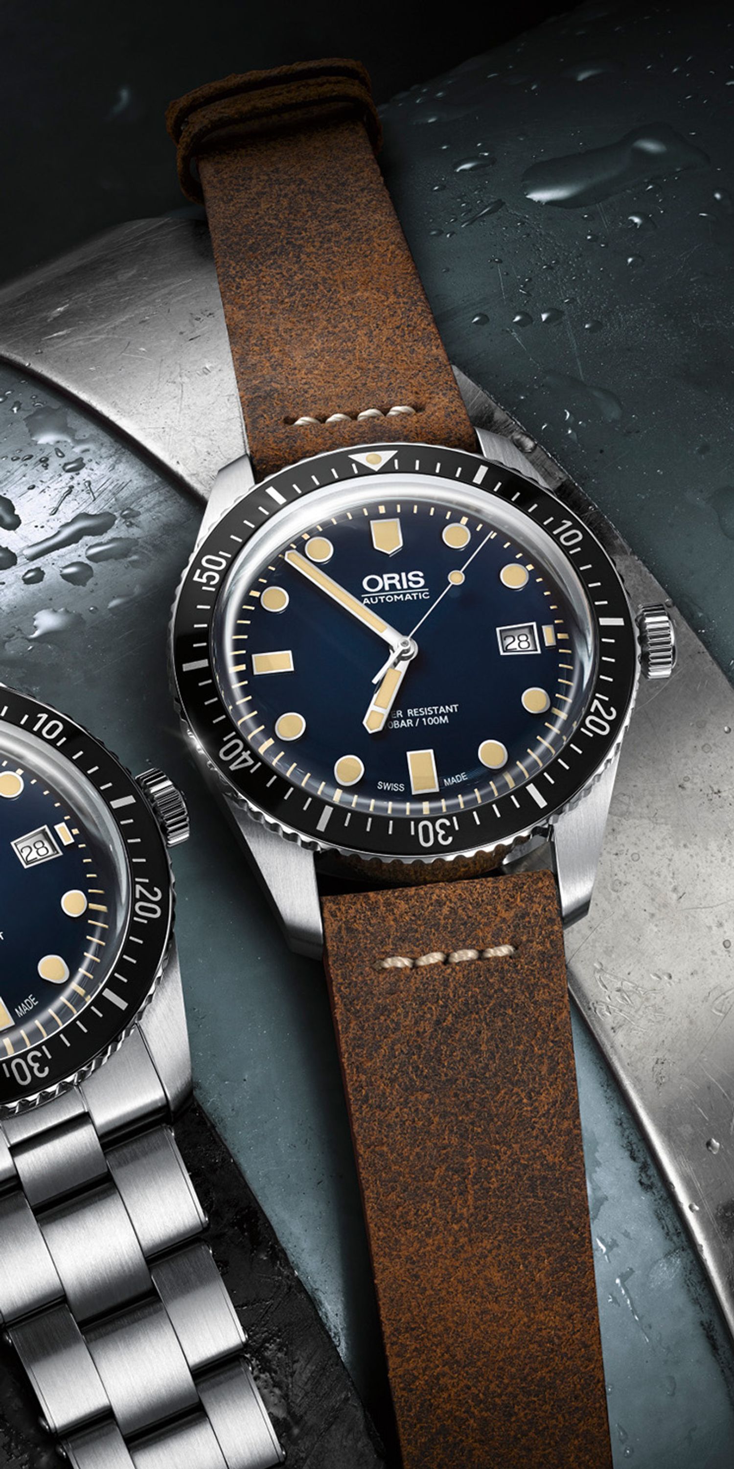 Close-up of the Oris Divers case with leather strap and dark blue dial. The indices and hands are bright yellow, and there is a date display at 3 o'clock. The silver case is complemented by a black bezel. To the left of the watch is another of the same model. Both are lying on a stone slab with water droplets visible in the upper section.