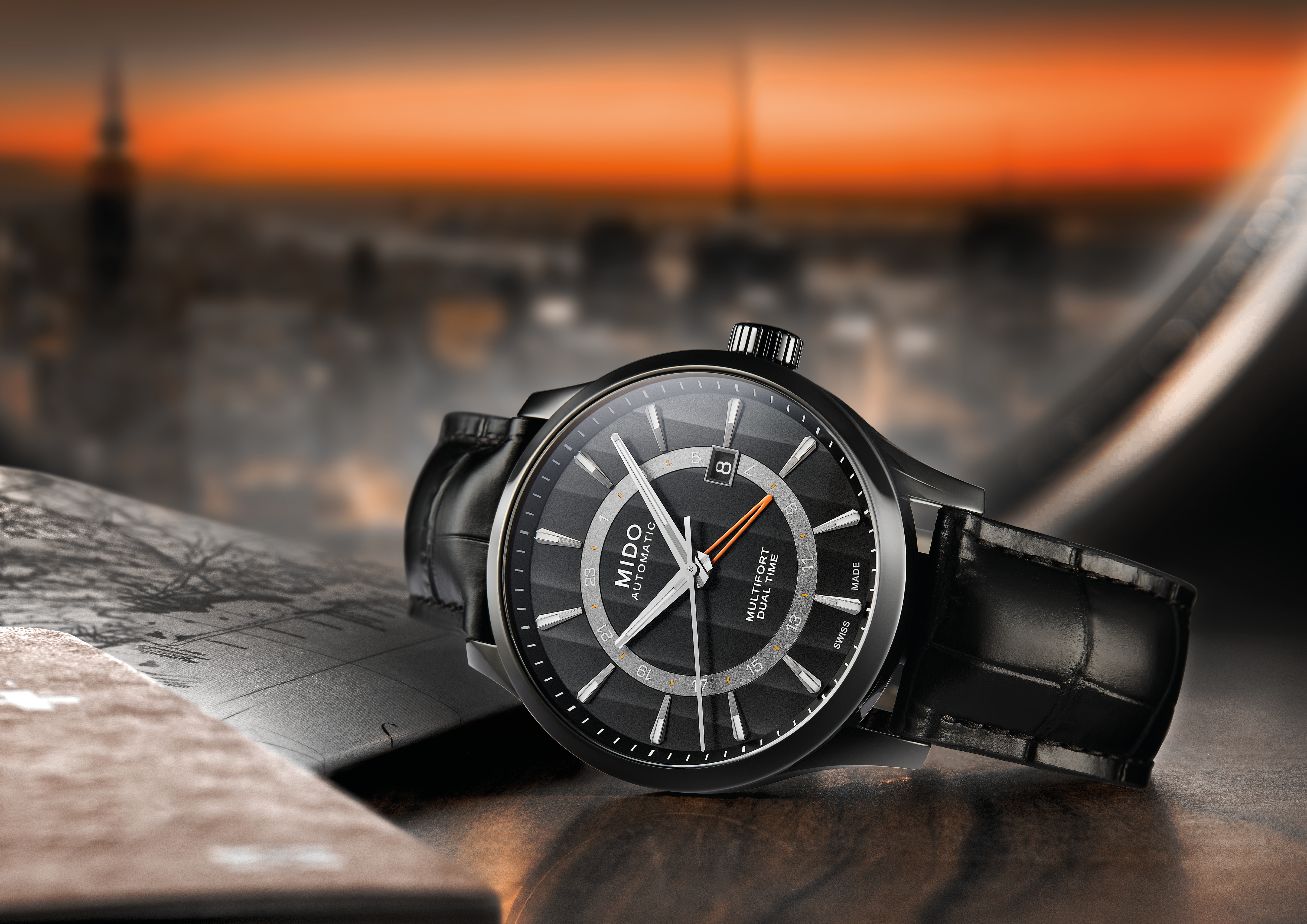 A shot of a Mido Multifort with a black leather strap and dial, featuring two time displays. In the background, the silhouette of a city can be seen.