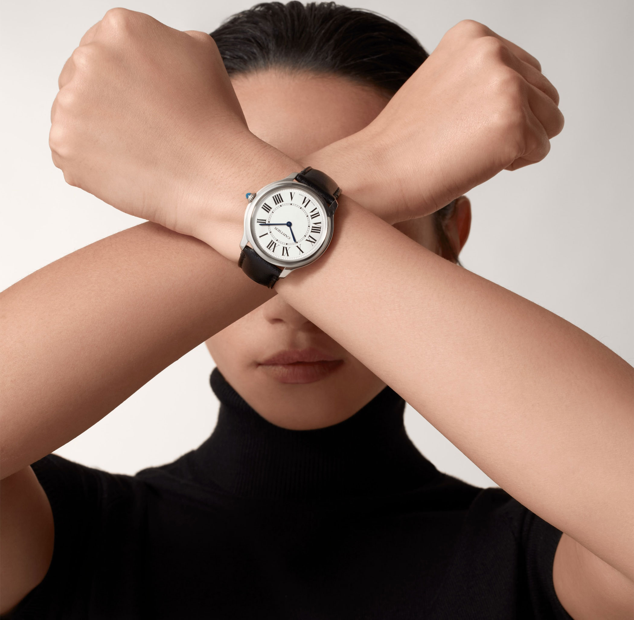 Ronde de Cartier with white dial, Roman numerals and black leather strap on a woman’s wrist, with her arms crossed in front of her face.