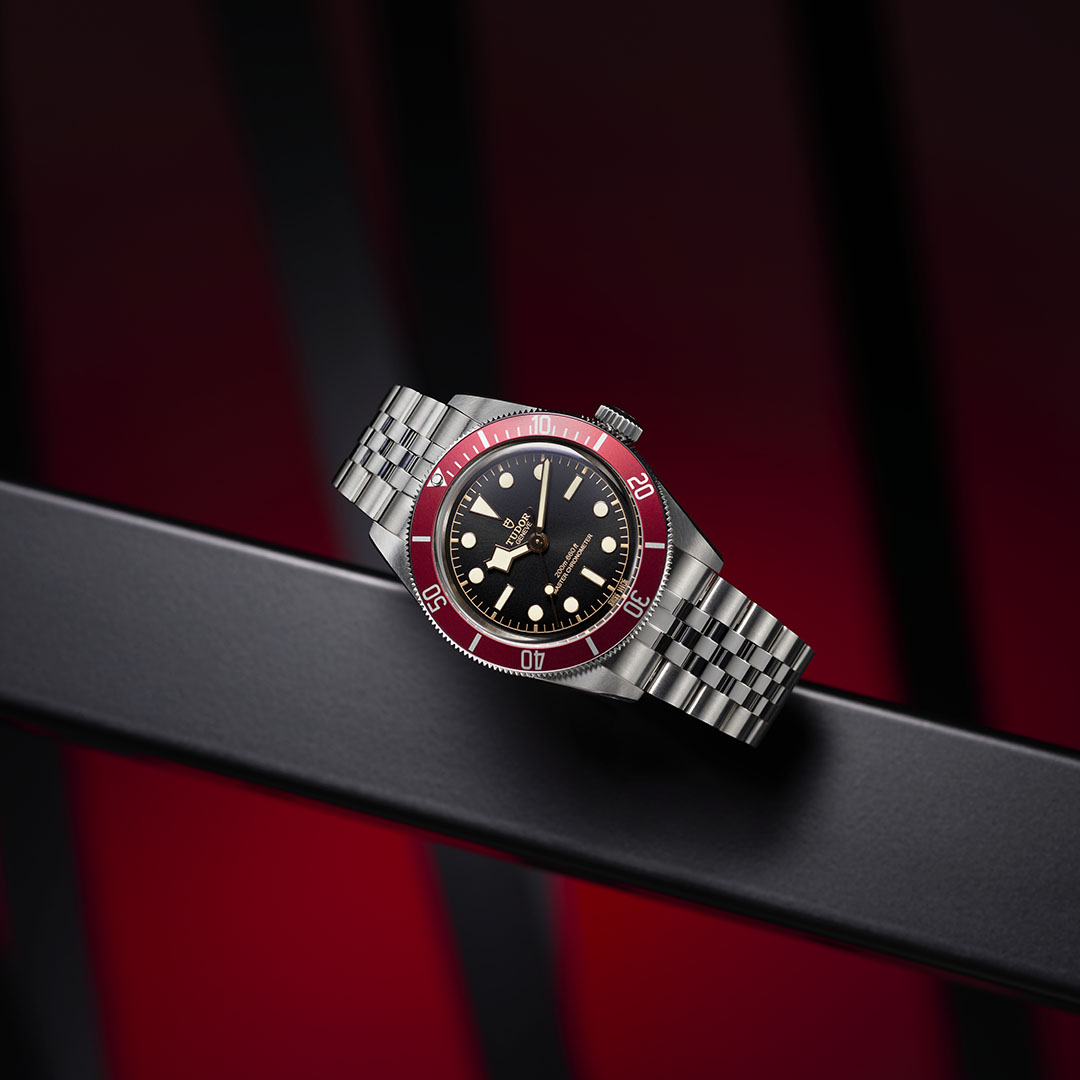 Nærbillede af Tudor GMT-uret med sort urskive, cremefarvede timemarkeringer, en rød og blå 'Pepsi'-krans og et urkasse i rustfrit stål. Baggrunden har dynamiske metalliske og røde accenter, som komplementerer urets dristige, funktionelle design og Tudors håndværkstradition.