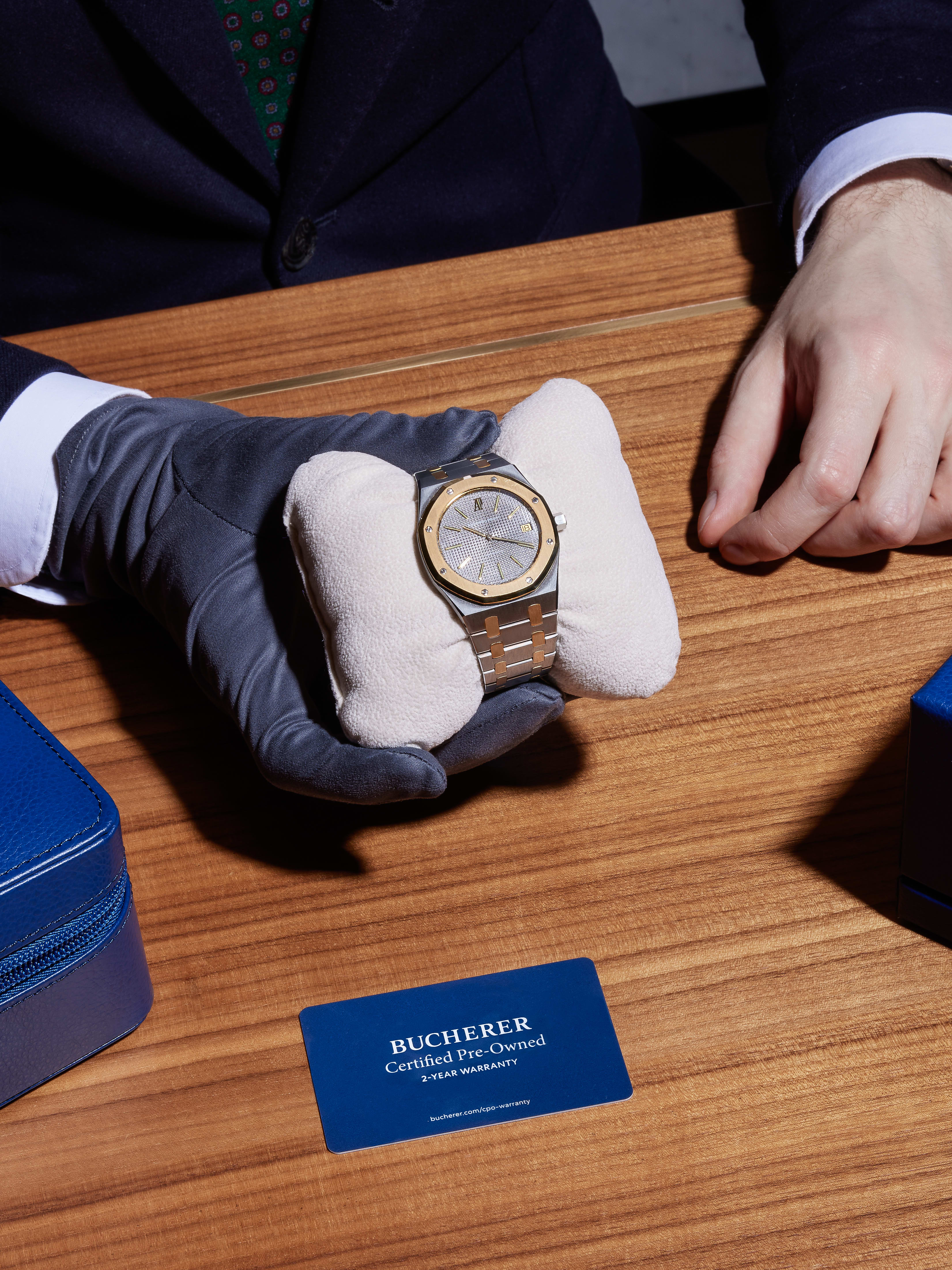 Eine behandschuhte Hand präsentiert eine silber-goldene Uhr auf einem weißen Kissen, daneben liegt eine blaue Bucherer-Karte auf einem Holztisch.