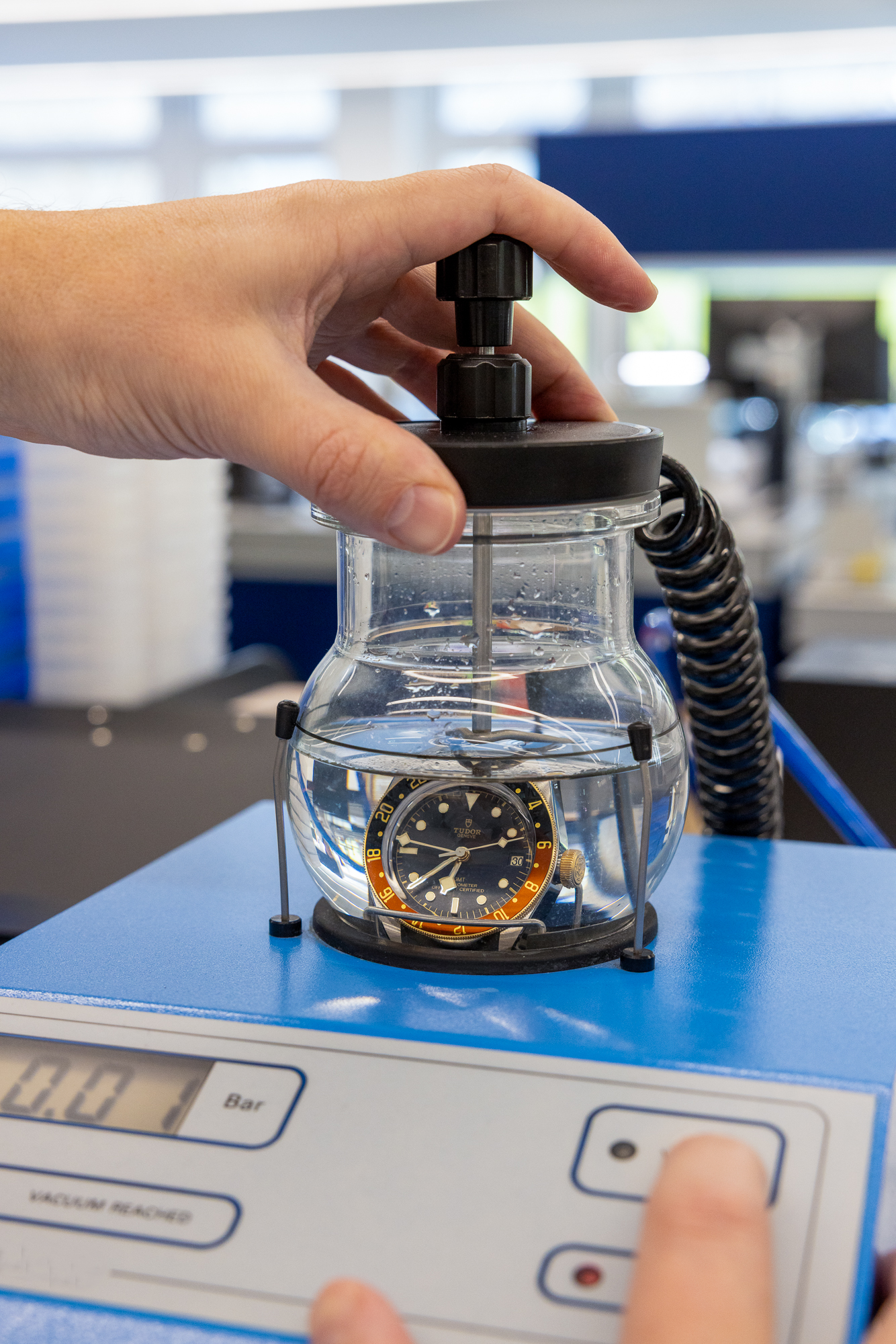 A hand operates a pump on a clear glass container with a watch submerged in water, on a blue machine.
