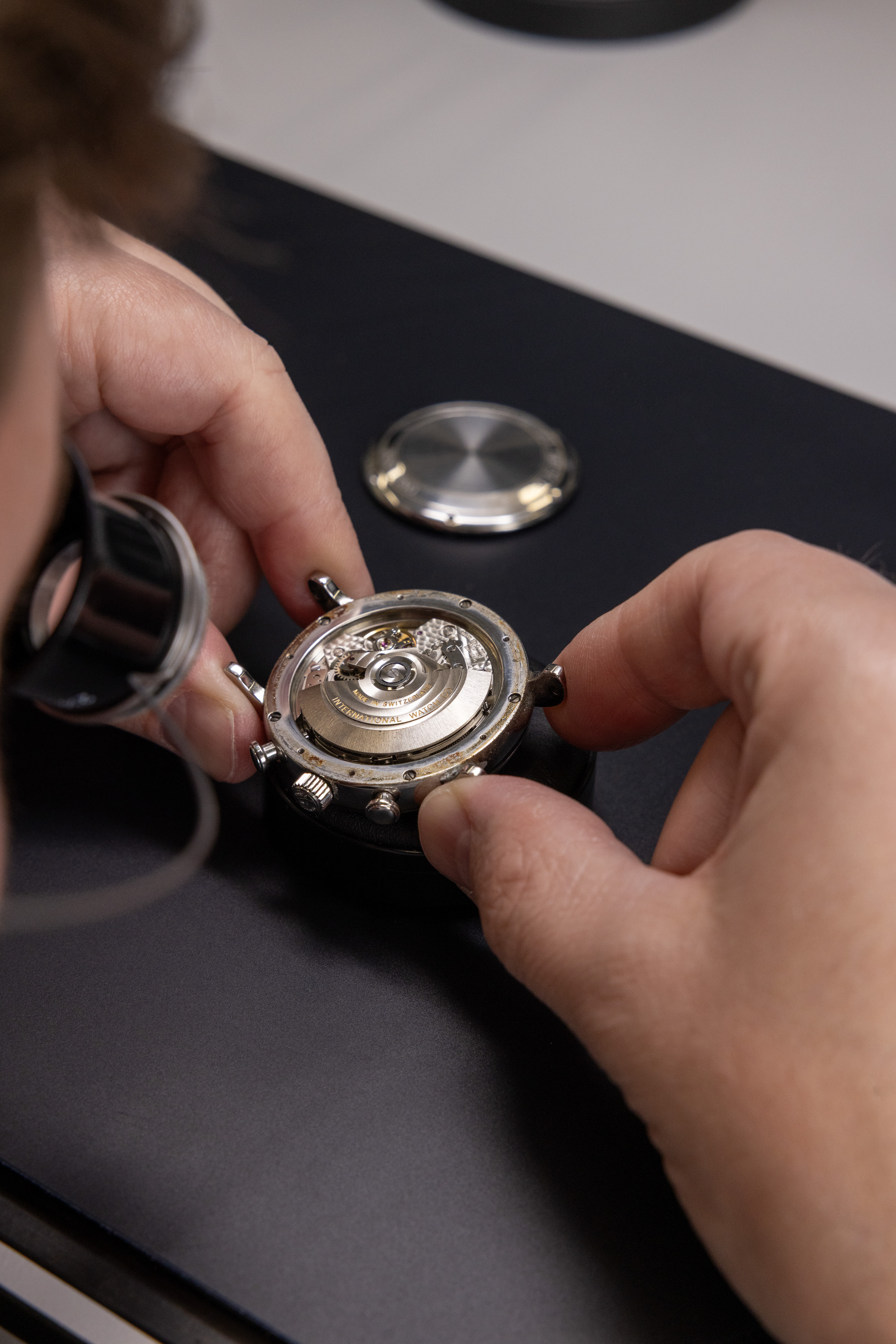 A watchmaker's hands hold an open watch, revealing its intricate mechanical movement, with a jeweler's loupe nearby.