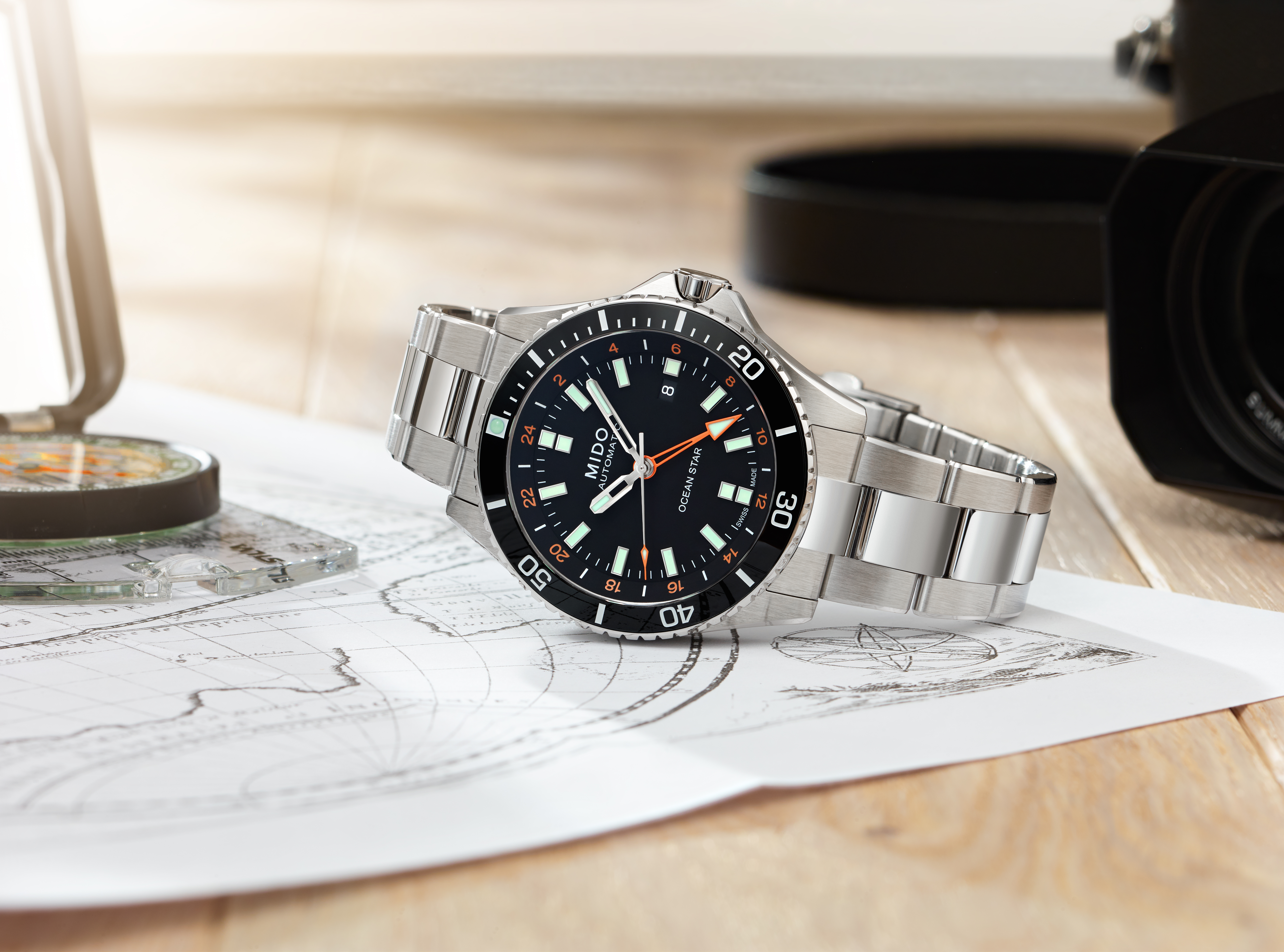 A silver Mido Ocean Star watch with a black face and bezel rests on a map next to a compass and camera.