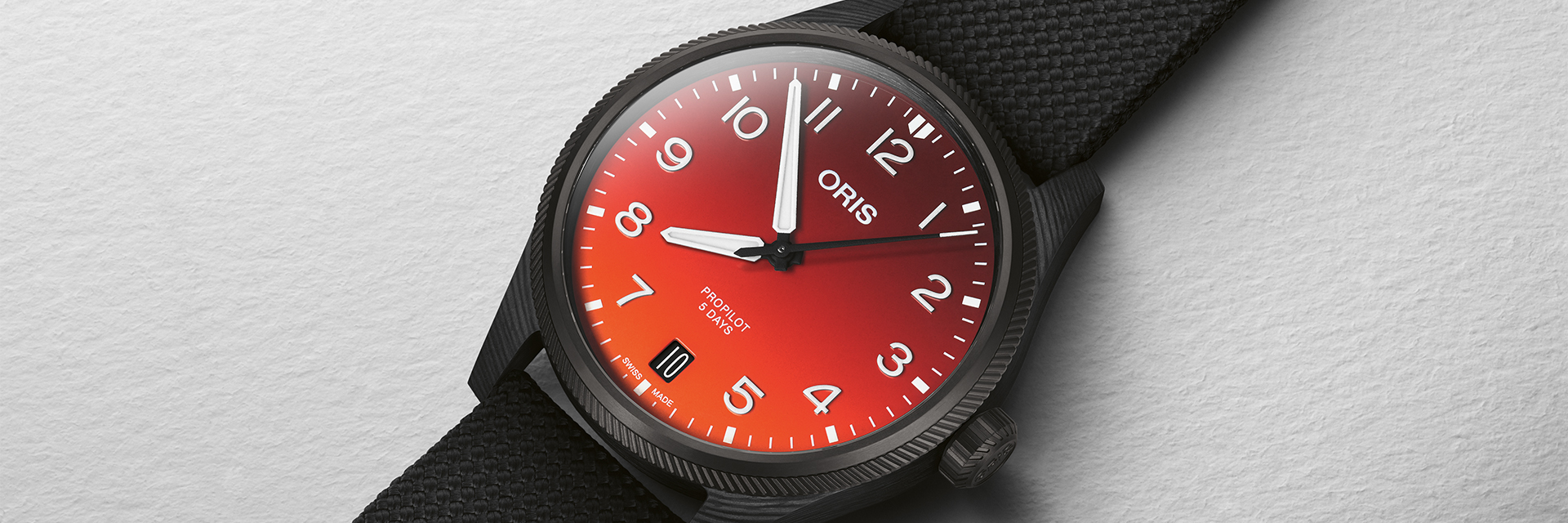  Close-up of an Oris Propilot Date with red dial and date display at 6 o'clock on a textile strap. The Oris logo is shown in the center of the lower edge of the image.