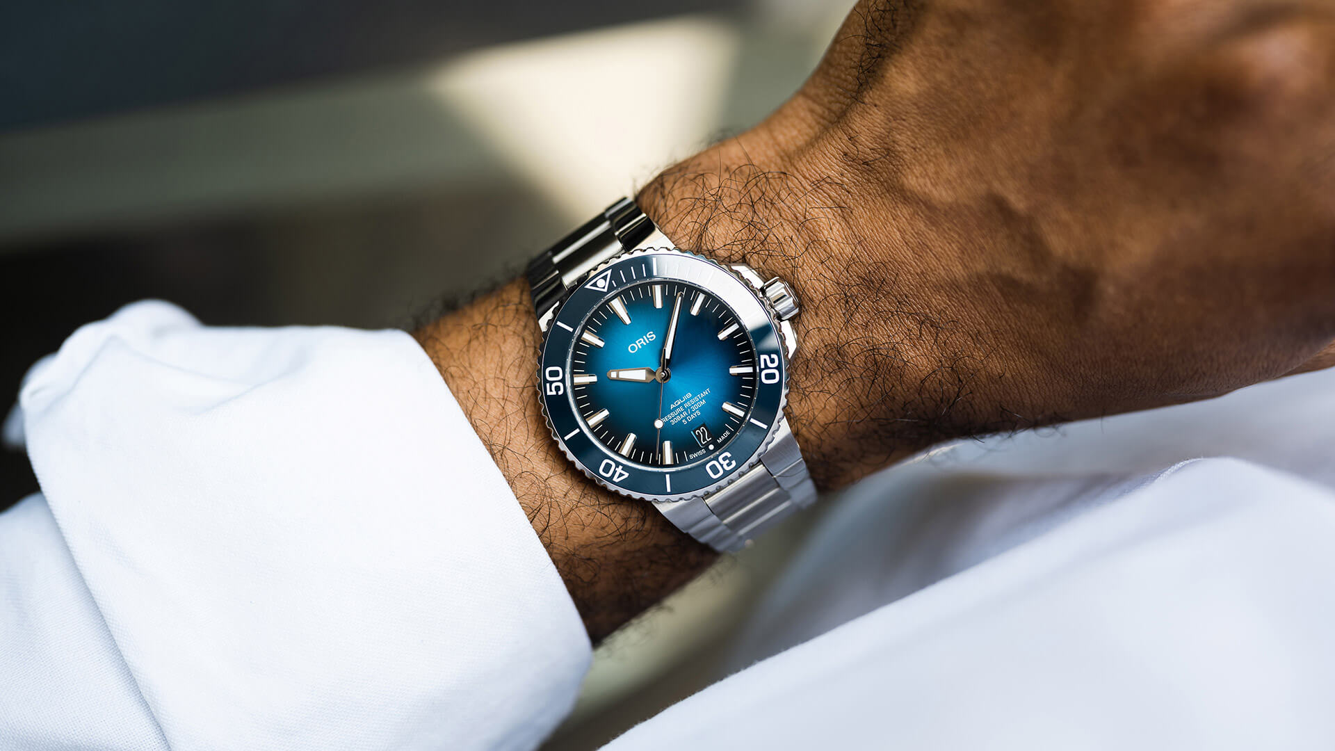 An Oris Aquis Date on a steel bracelet on the wrist of a man wearing a white shirt. The dial and bezel of the watch are blue.