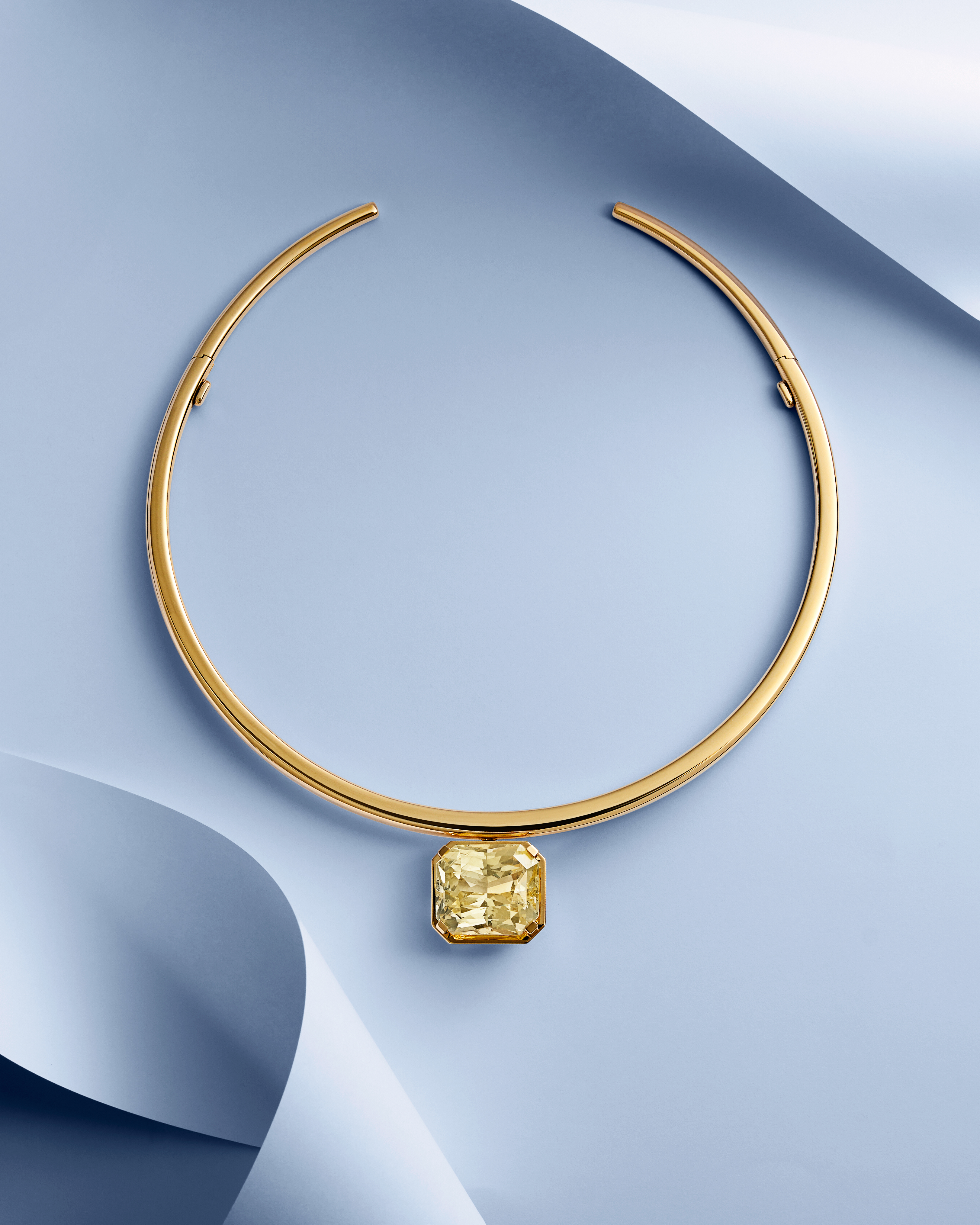 A gold choker necklace with a large, rectangular yellow diamond pendant, on a light blue background with curved paper elements.