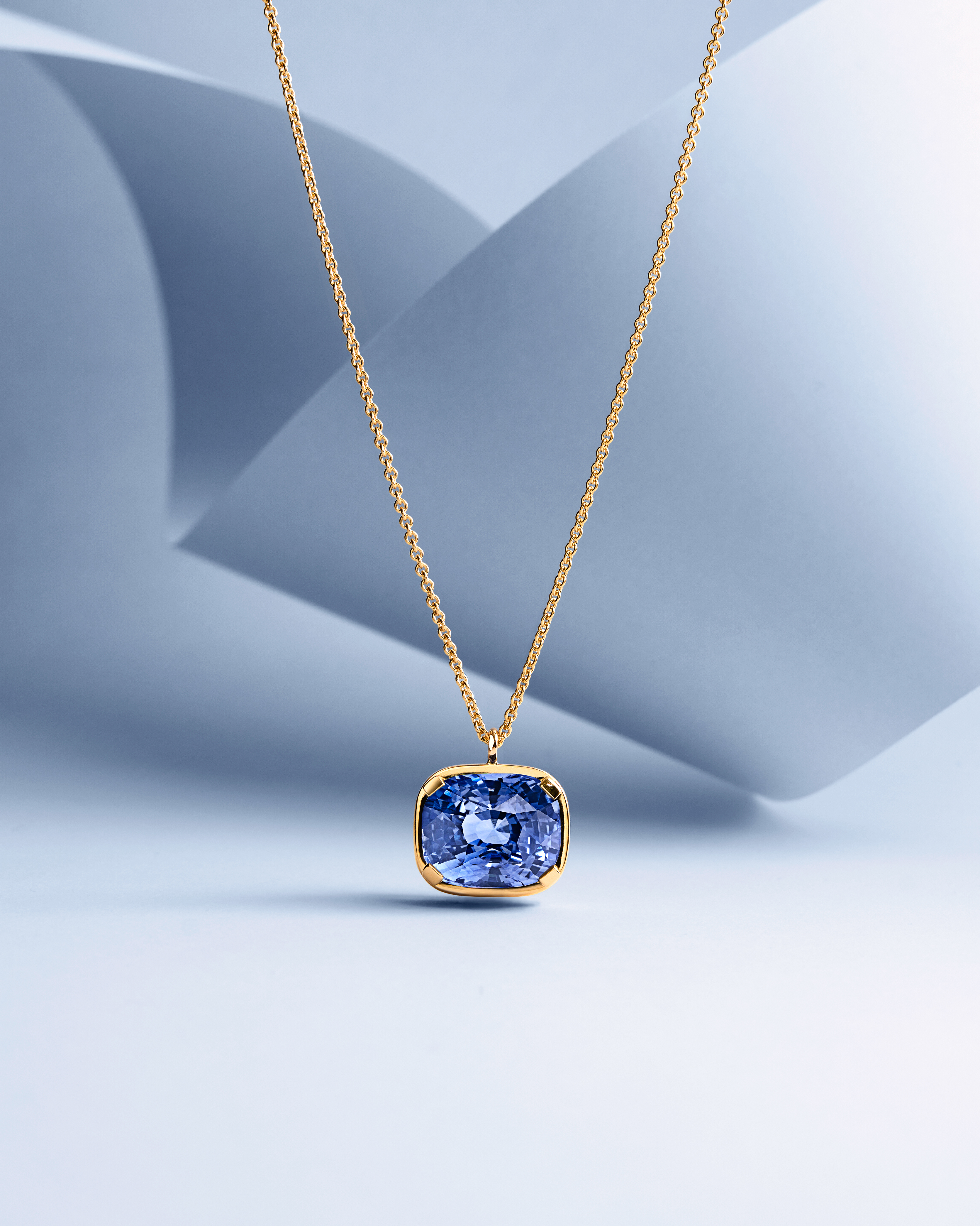 A gold chain necklace with a cushion-cut blue sapphire pendant against a light blue and white folded paper background.