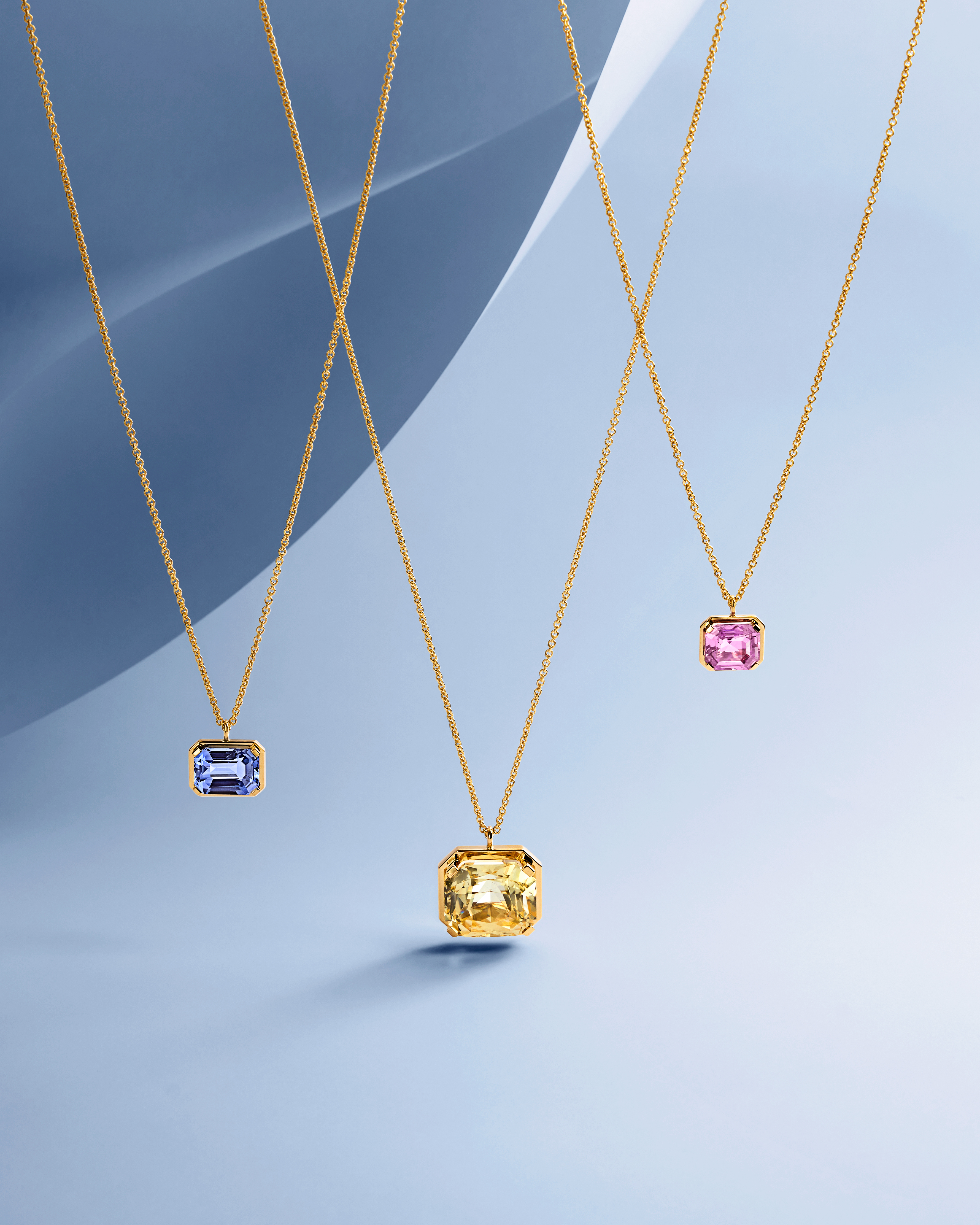 Three gold necklaces with rectangular gemstone pendants: blue, yellow, and pink, against a light blue background.
