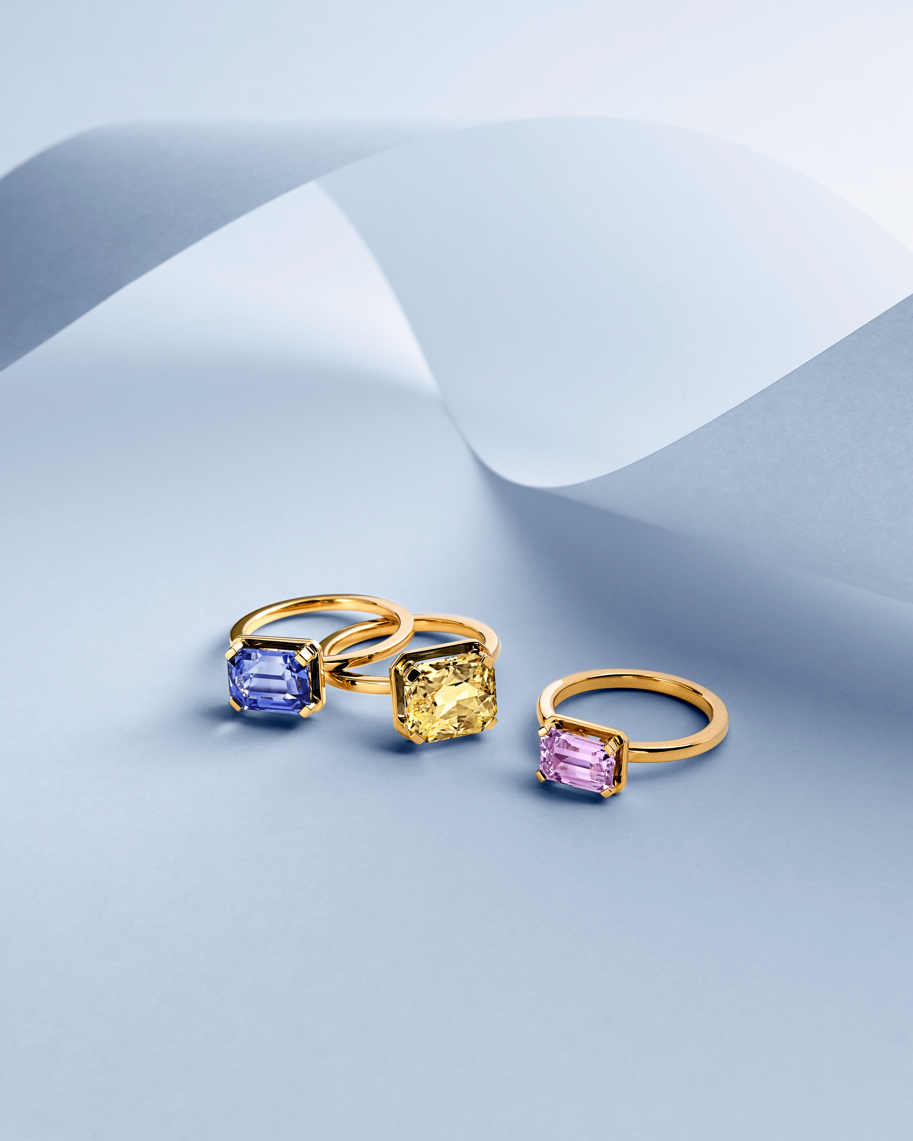 Three gold rings with emerald-cut gemstones: blue, yellow, and pink, on a light blue surface with curved paper.