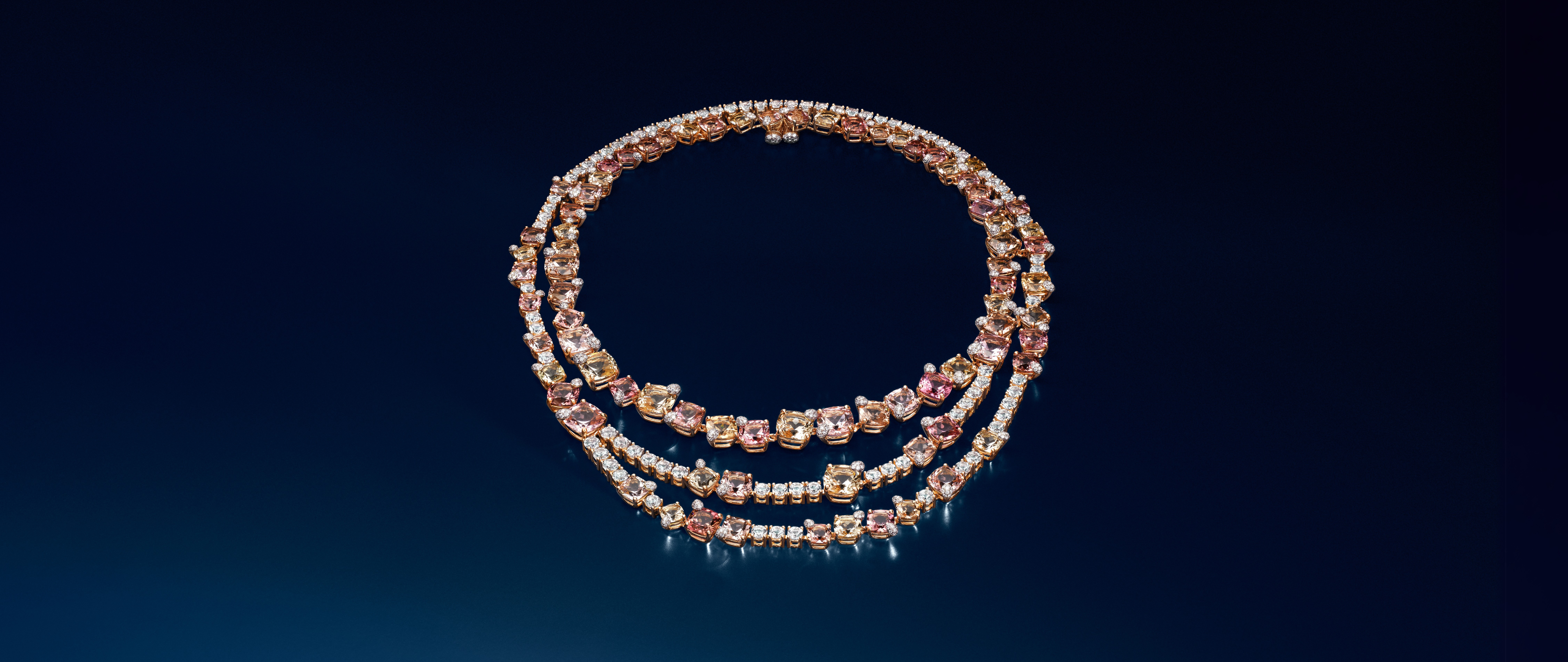 Three layered necklaces with square-cut pink, yellow, and clear gemstones on a dark blue background.