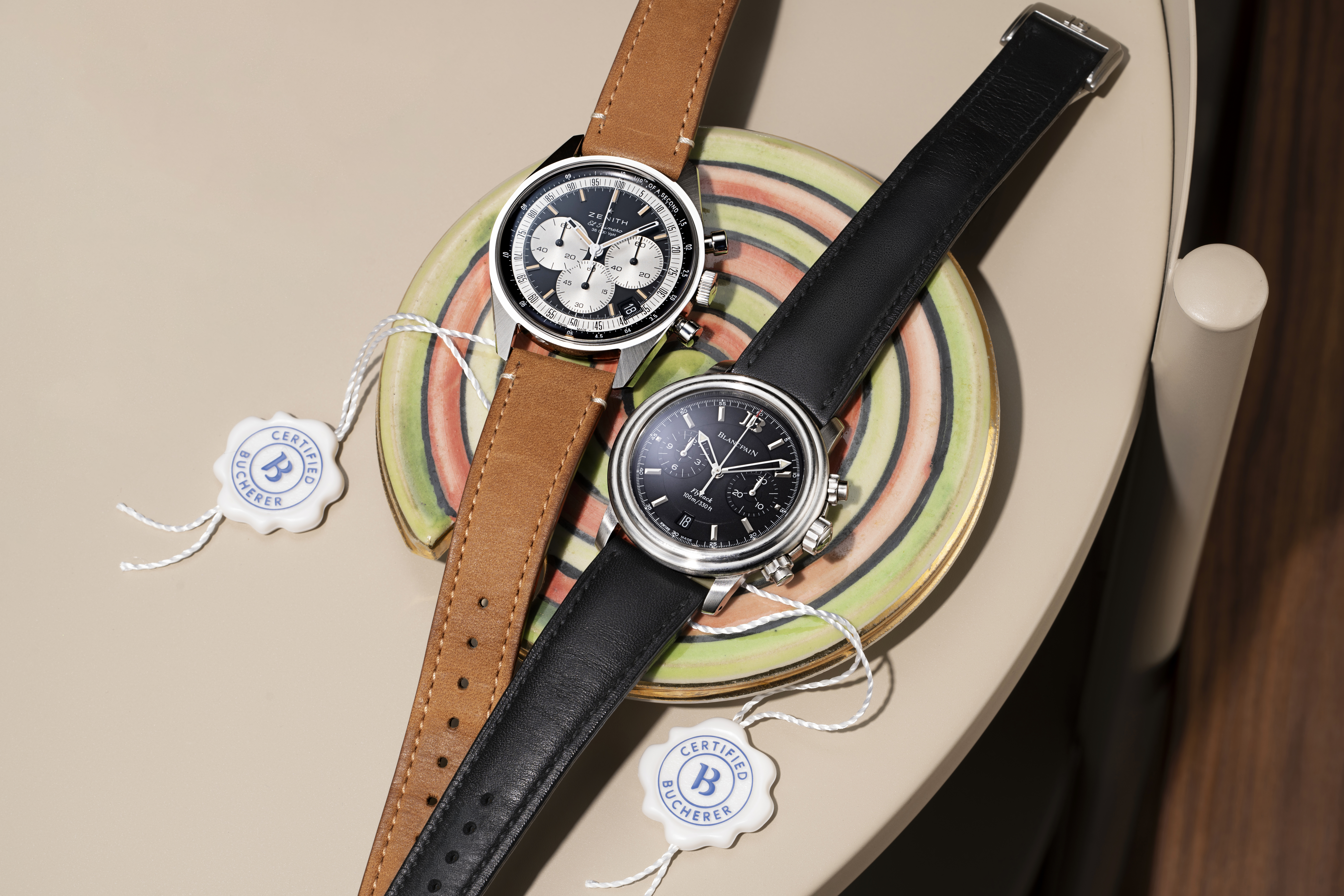 Two luxury chronographs, one with a brown leather strap and white dial, the other with a black leather strap and black dial, rest on a colorful plate.