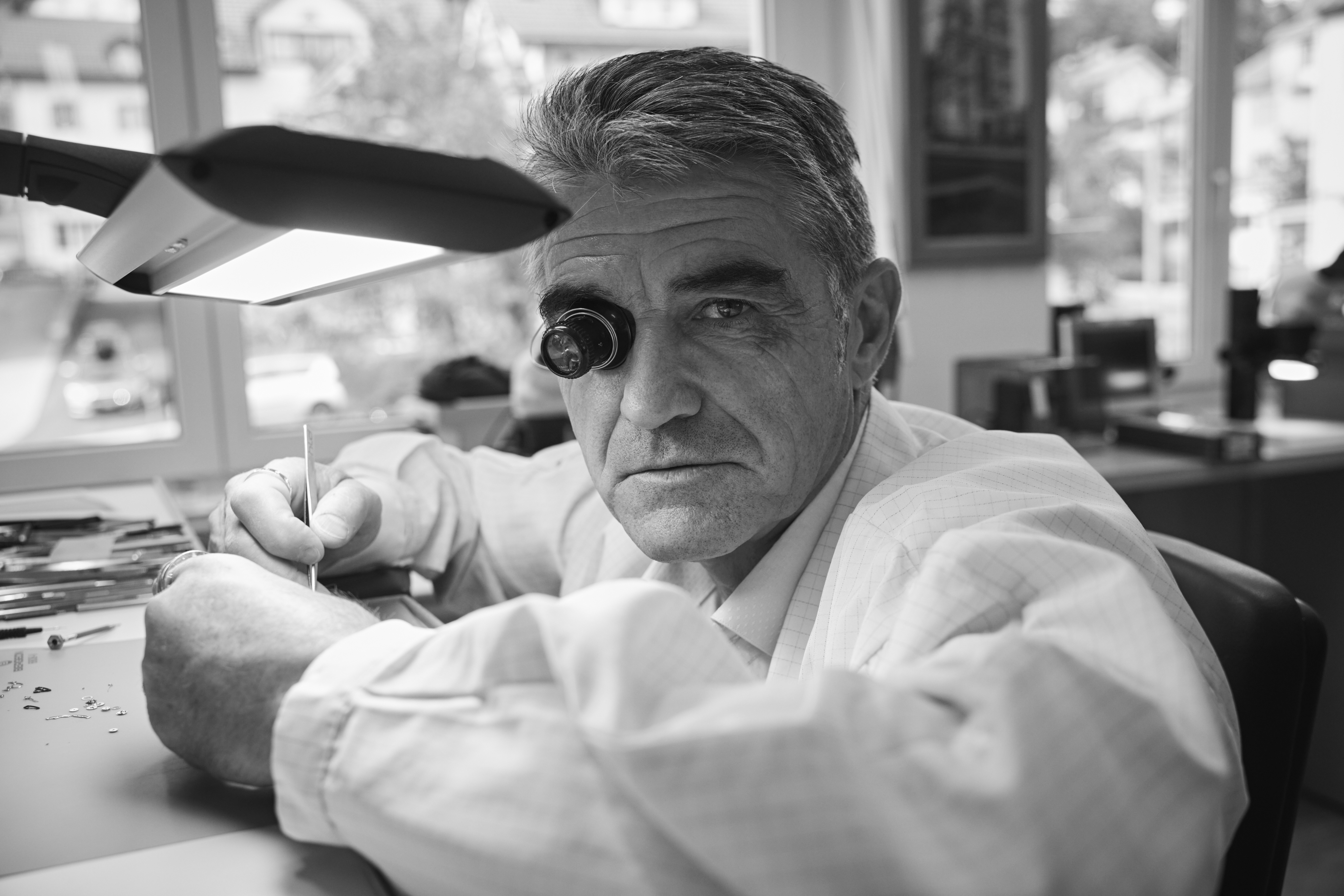 A man with a jeweler's loupe in his eye works on a small object under a desk lamp.
