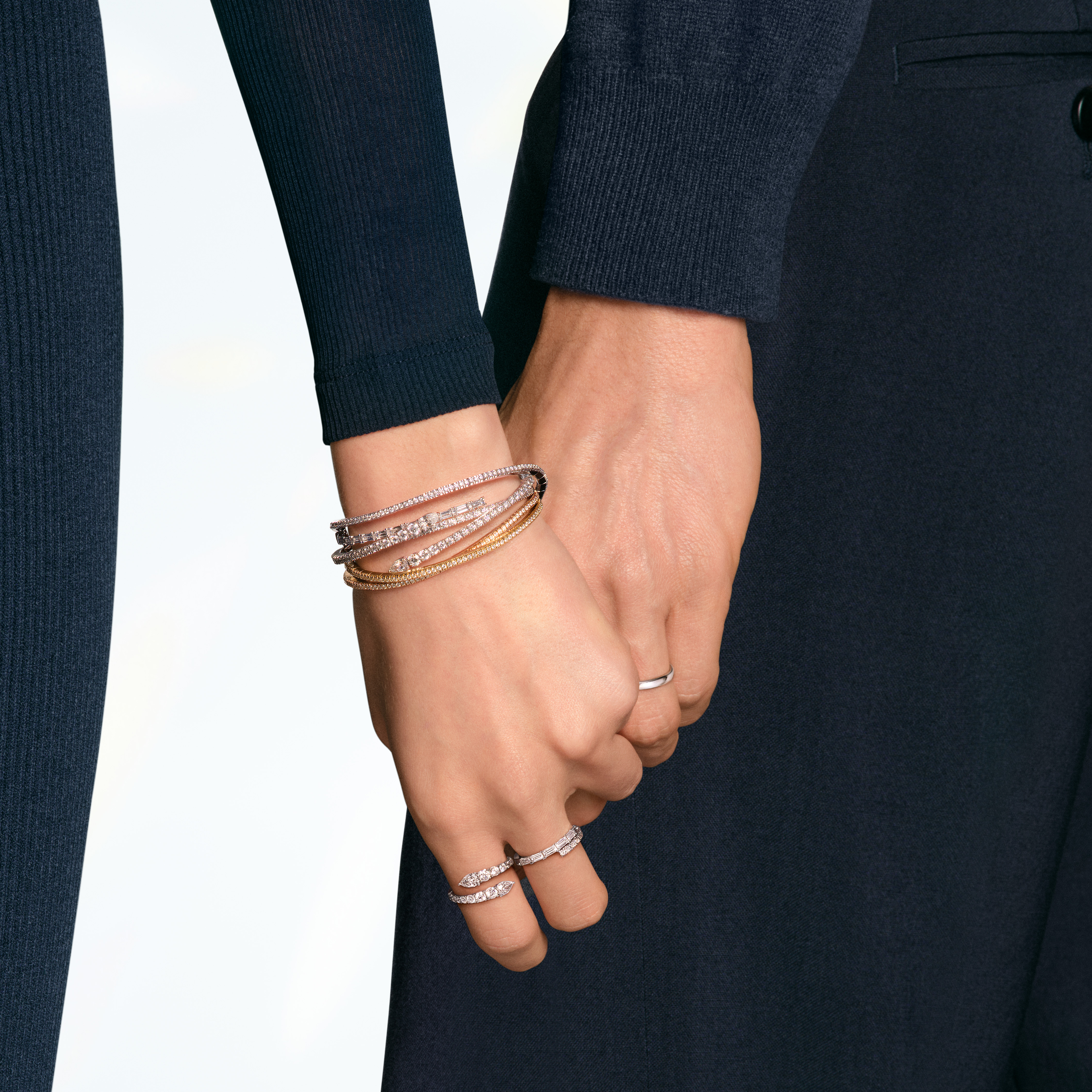 Two people holding hands, one wearing multiple diamond bracelets and rings, the other a simple silver band.
