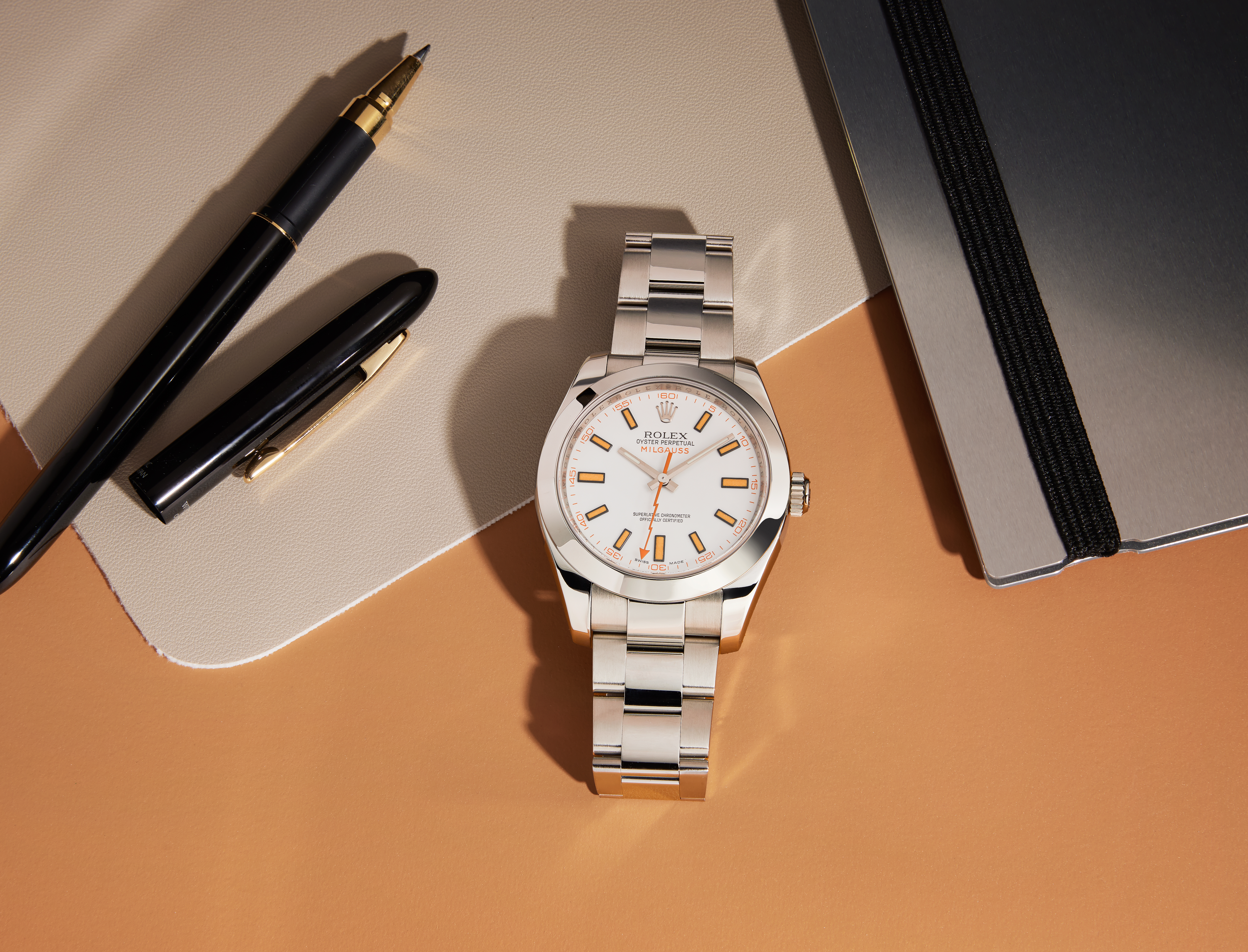 Eine silberne Rolex Milgauss Uhr mit weißem Zifferblatt und orangen Akzenten, ein schwarzer Stift und ein silbernes Notizbuch auf beigem und orangenem Untergrund.
