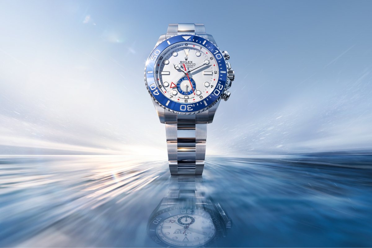 Une montre Rolex argentée, à cadran blanc et lunette bleue, se dresse sur une surface bleue réfléchissante sous un ciel lumineux.