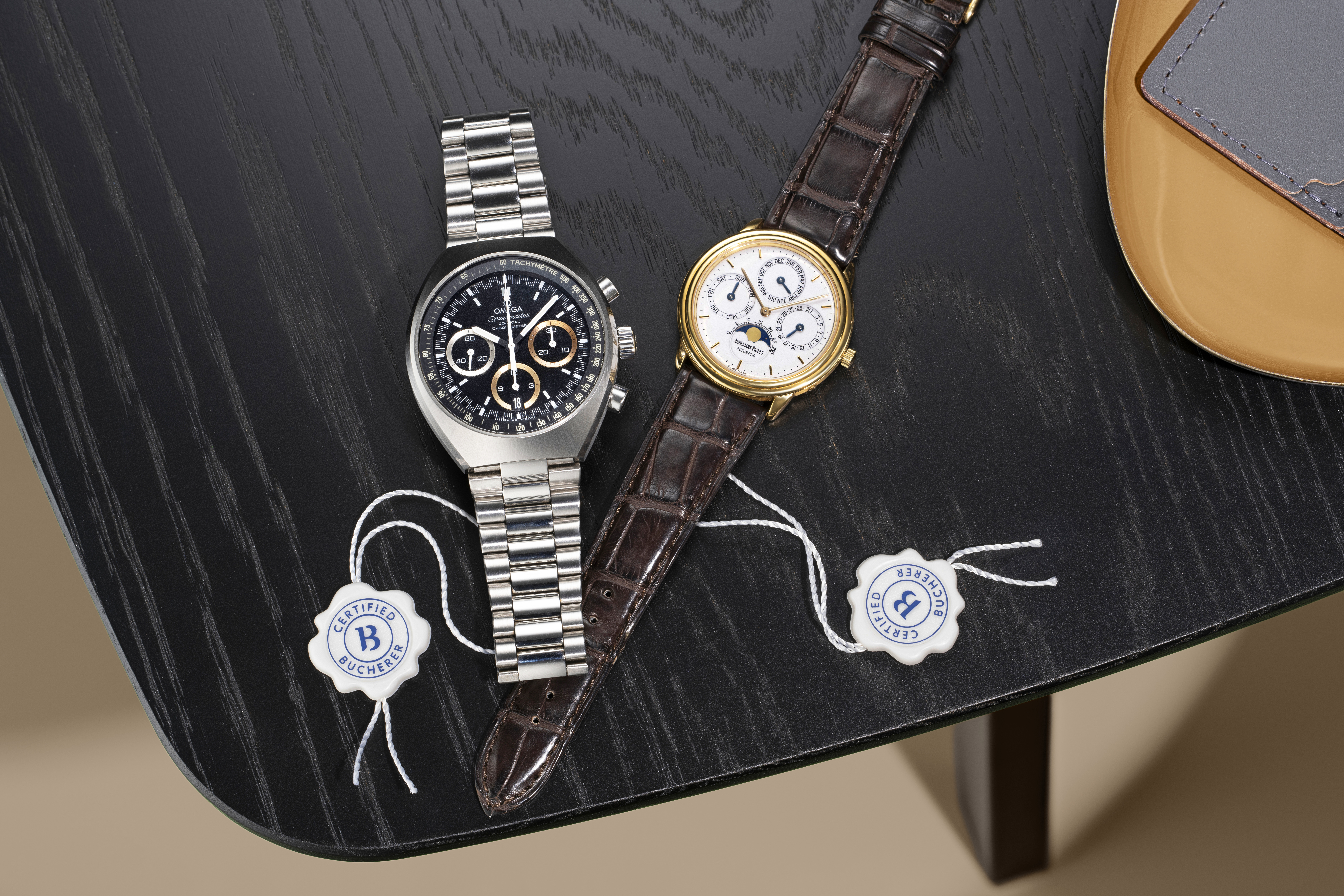 Deux montres de luxe, l'une avec un bracelet argenté et un cadran noir, l'autre avec un bracelet en cuir marron et un cadran blanc, posées sur une table en bois foncé.