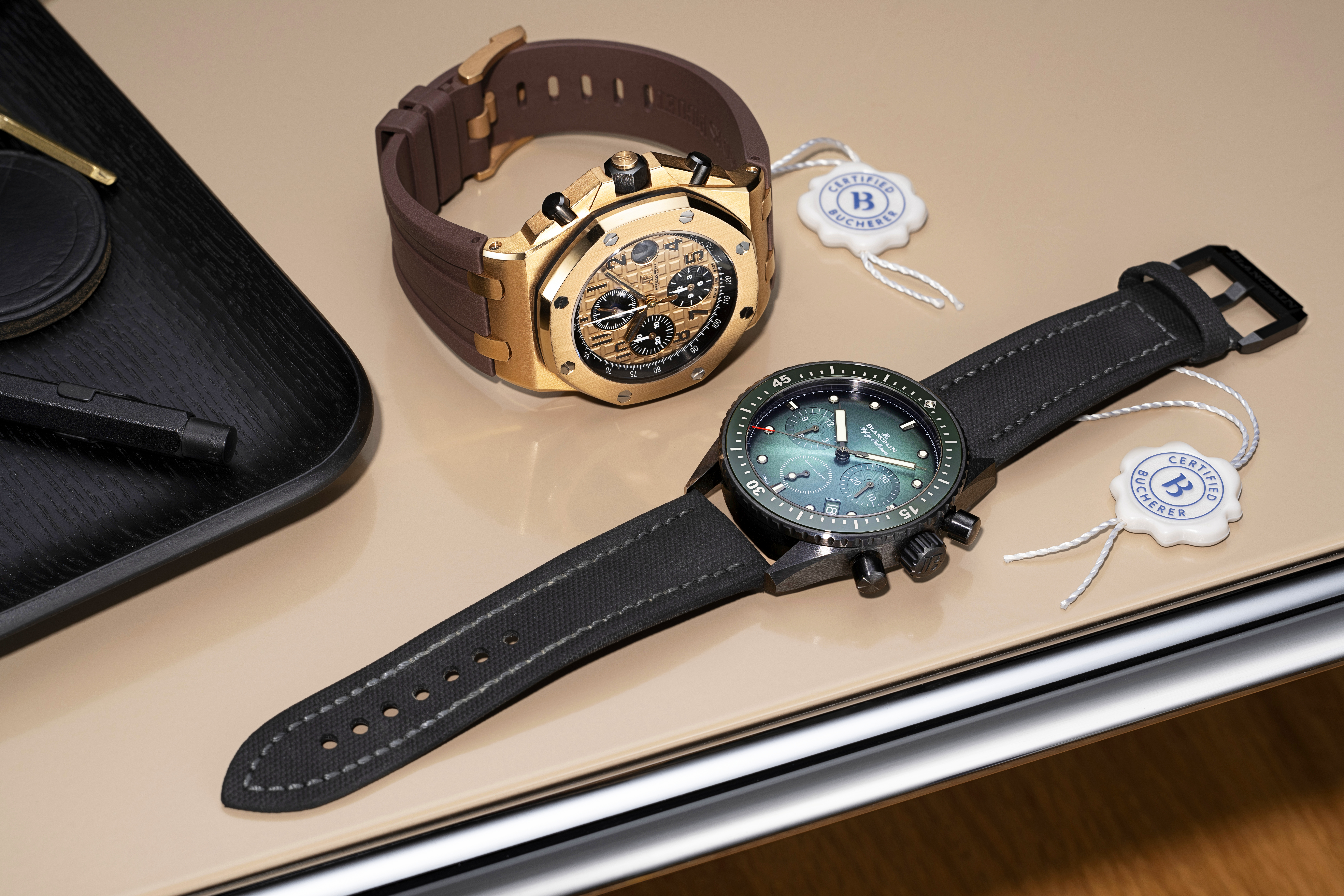 Two luxury wristwatches, one gold with a brown band and one black with a green face, on a beige surface.