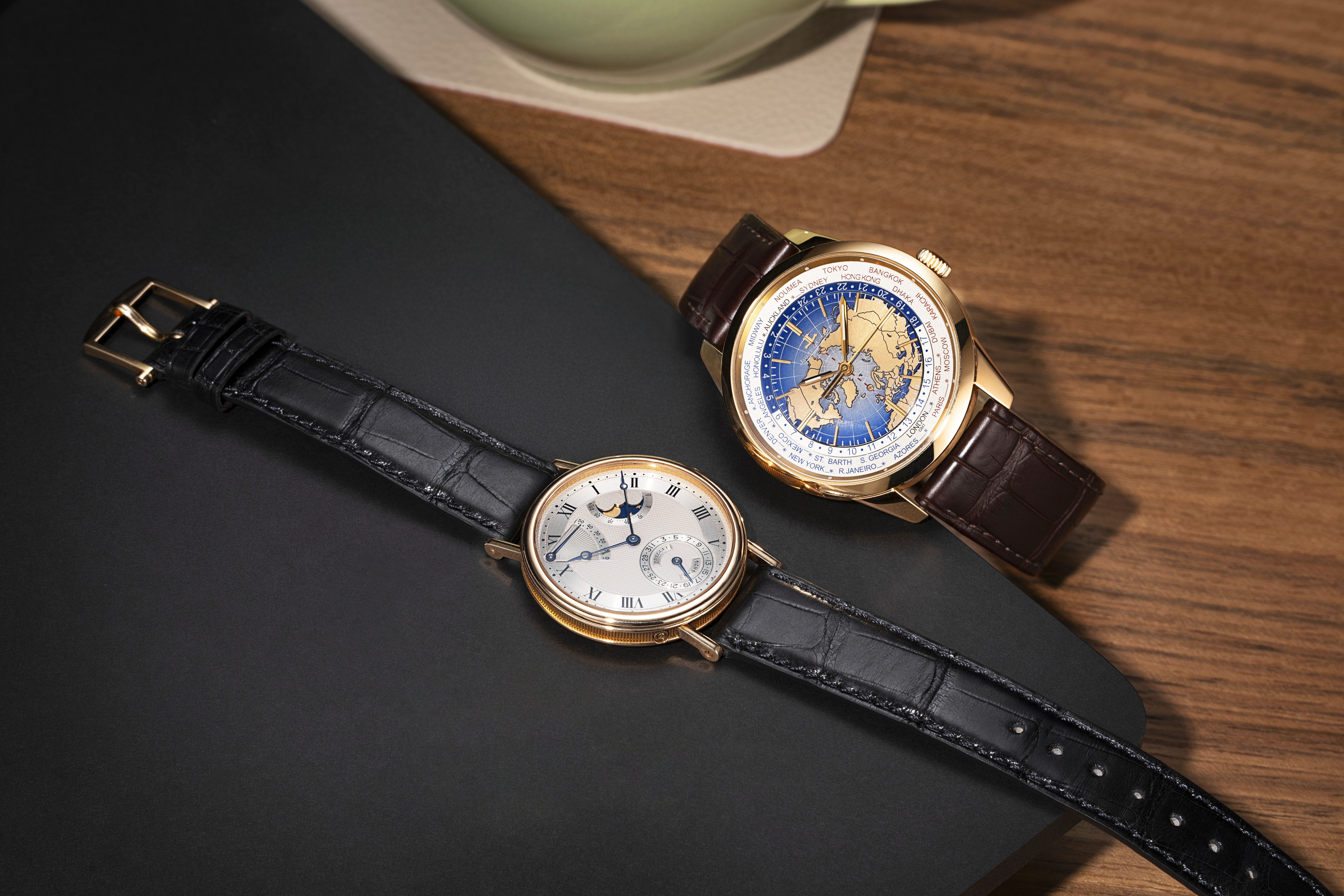 Two luxury wristwatches, one with a white face and black leather strap, the other with a blue world map face and brown leather strap.