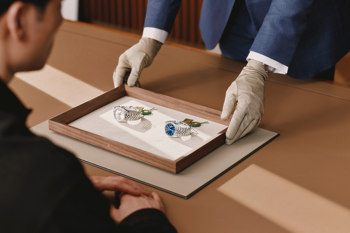 A person in gloves presents a wooden tray with two luxury watches to a customer.