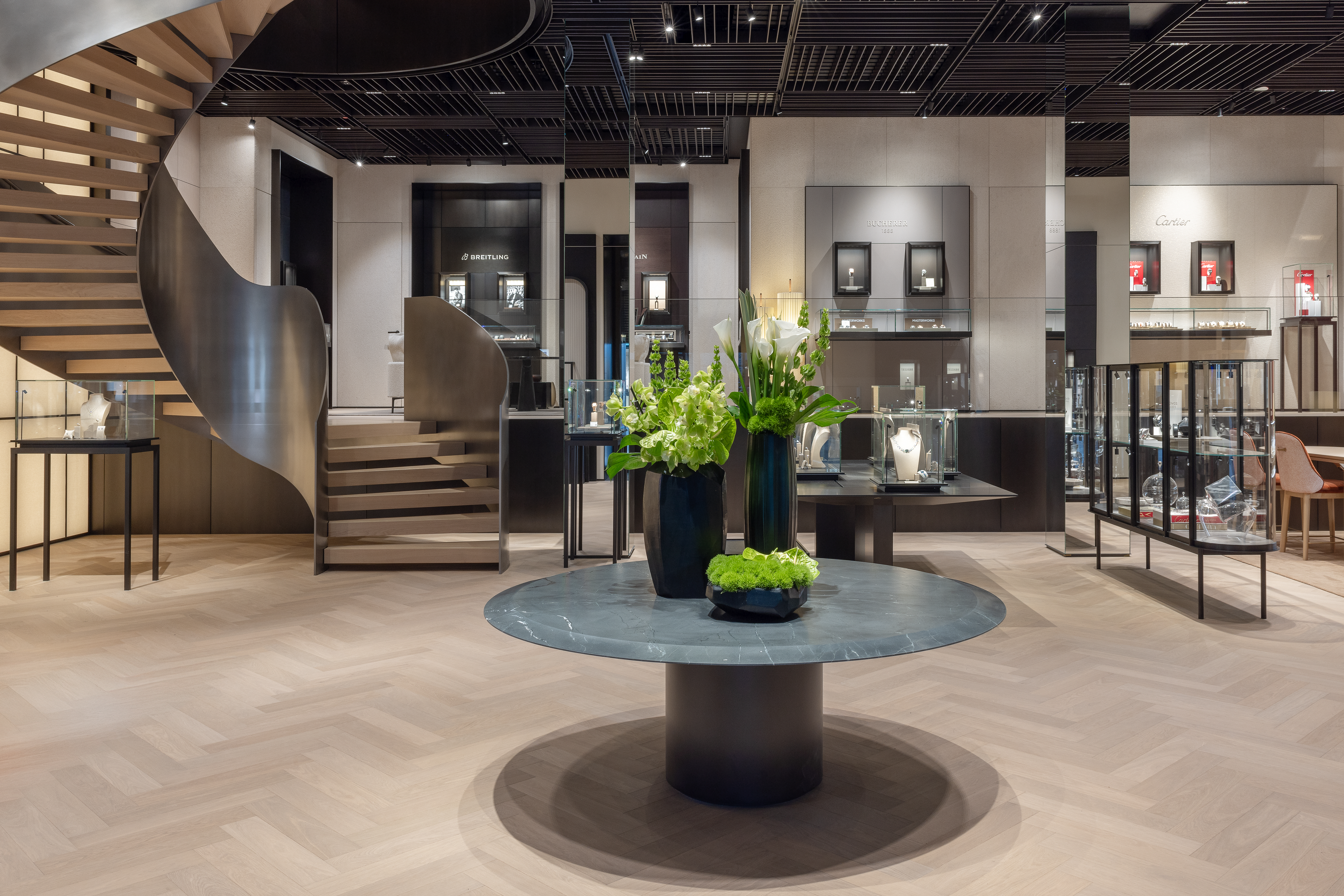 A luxurious retail store interior with a spiral staircase, display cases, and a round table with floral arrangements.