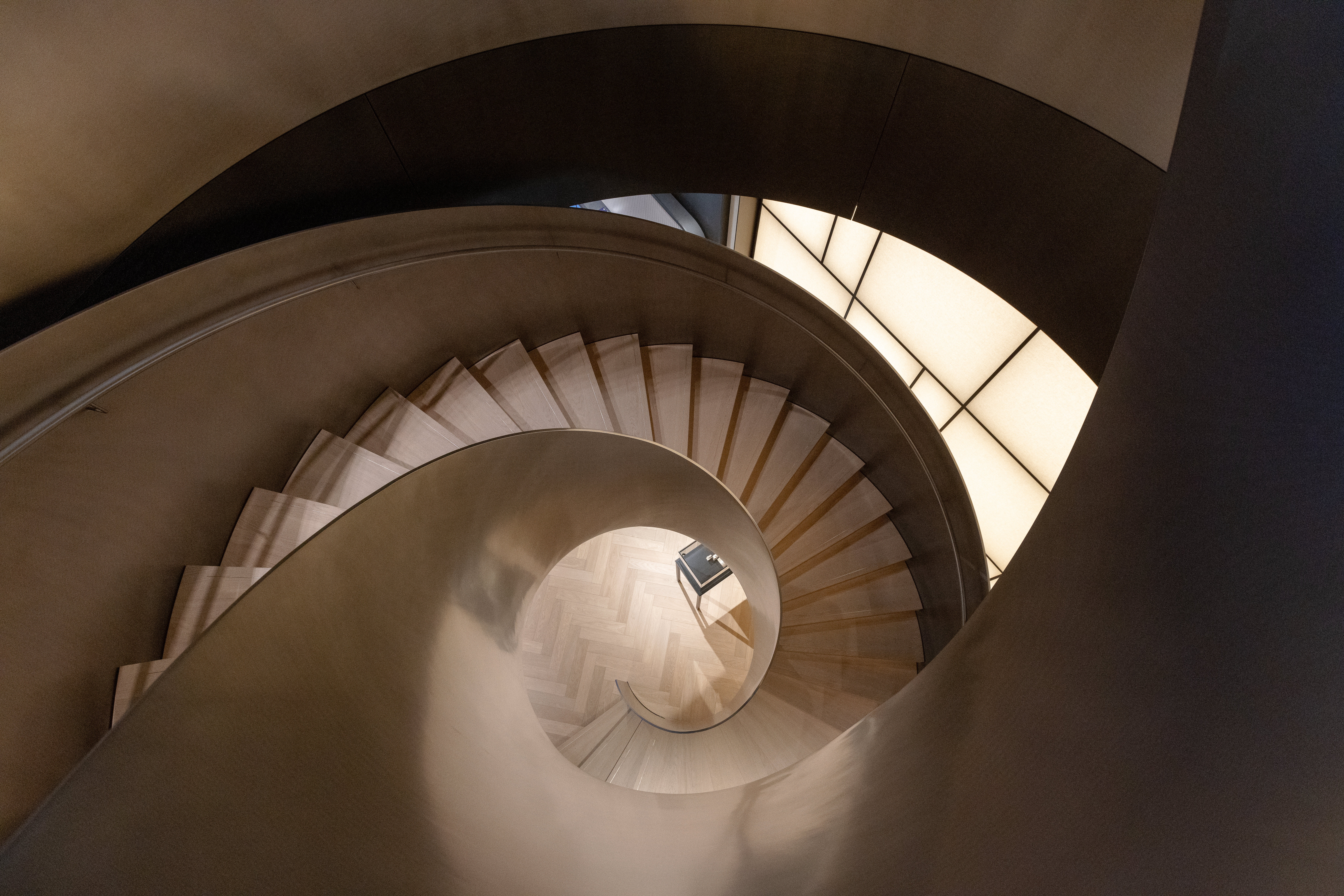Eine Wendeltreppe mit hellen Stufen und dunklem Geländer, von oben betrachtet.