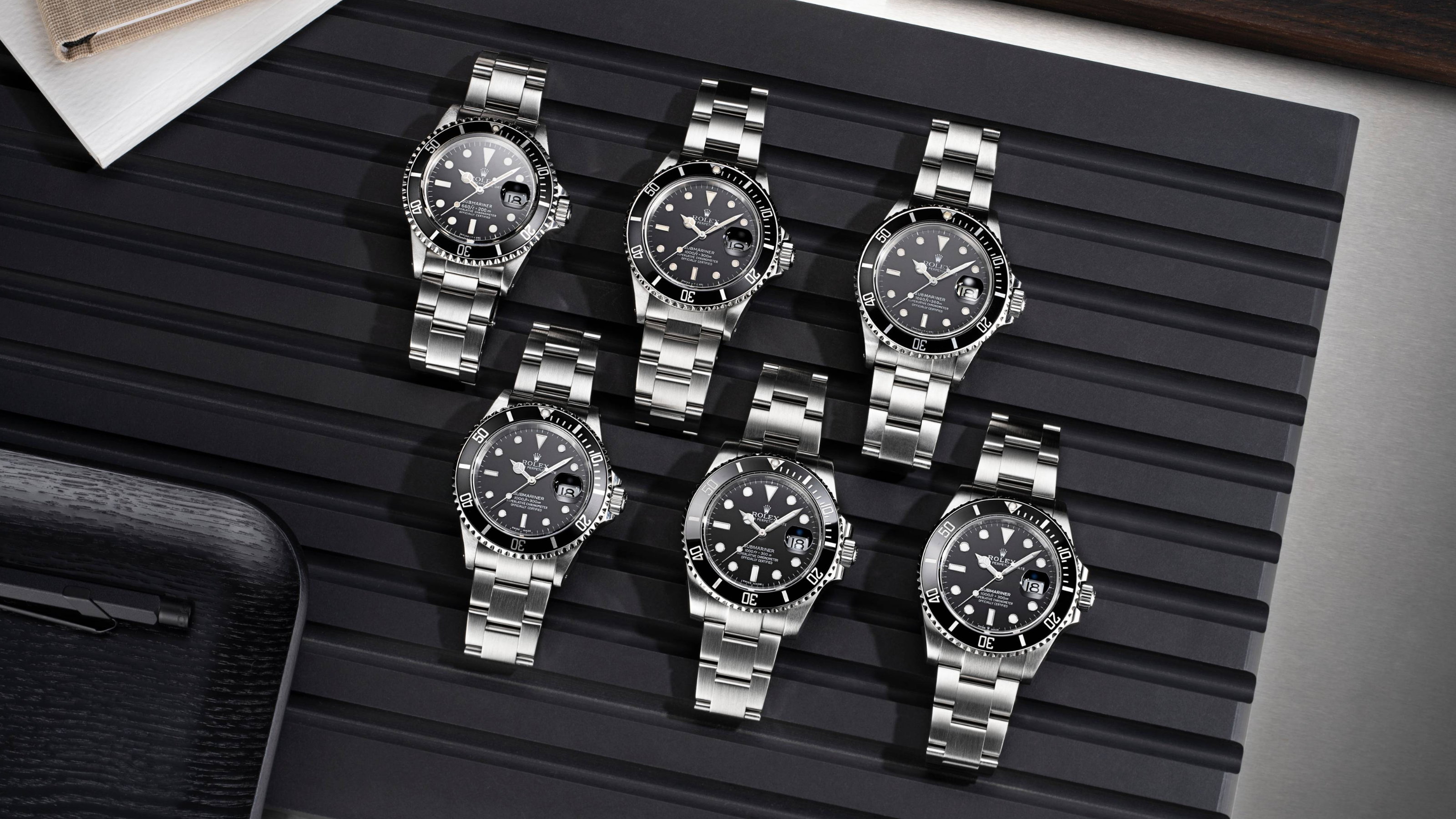 Six montres Rolex Submariner à cadrans noirs et bracelets en acier inoxydable, disposées sur une surface noire côtelée.