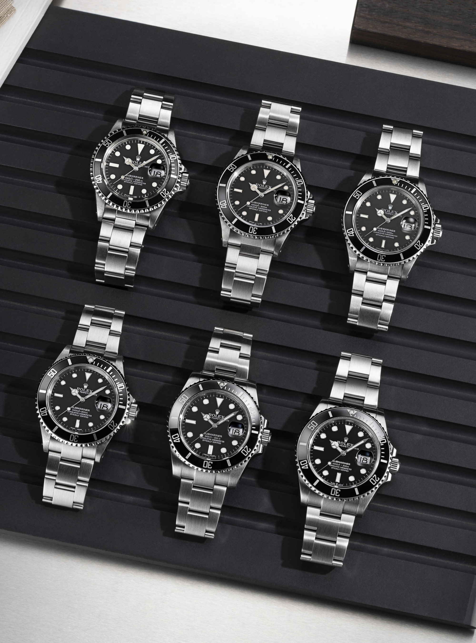 Six montres argentées à cadran et lunette noirs, disposées sur une surface noire striée.
