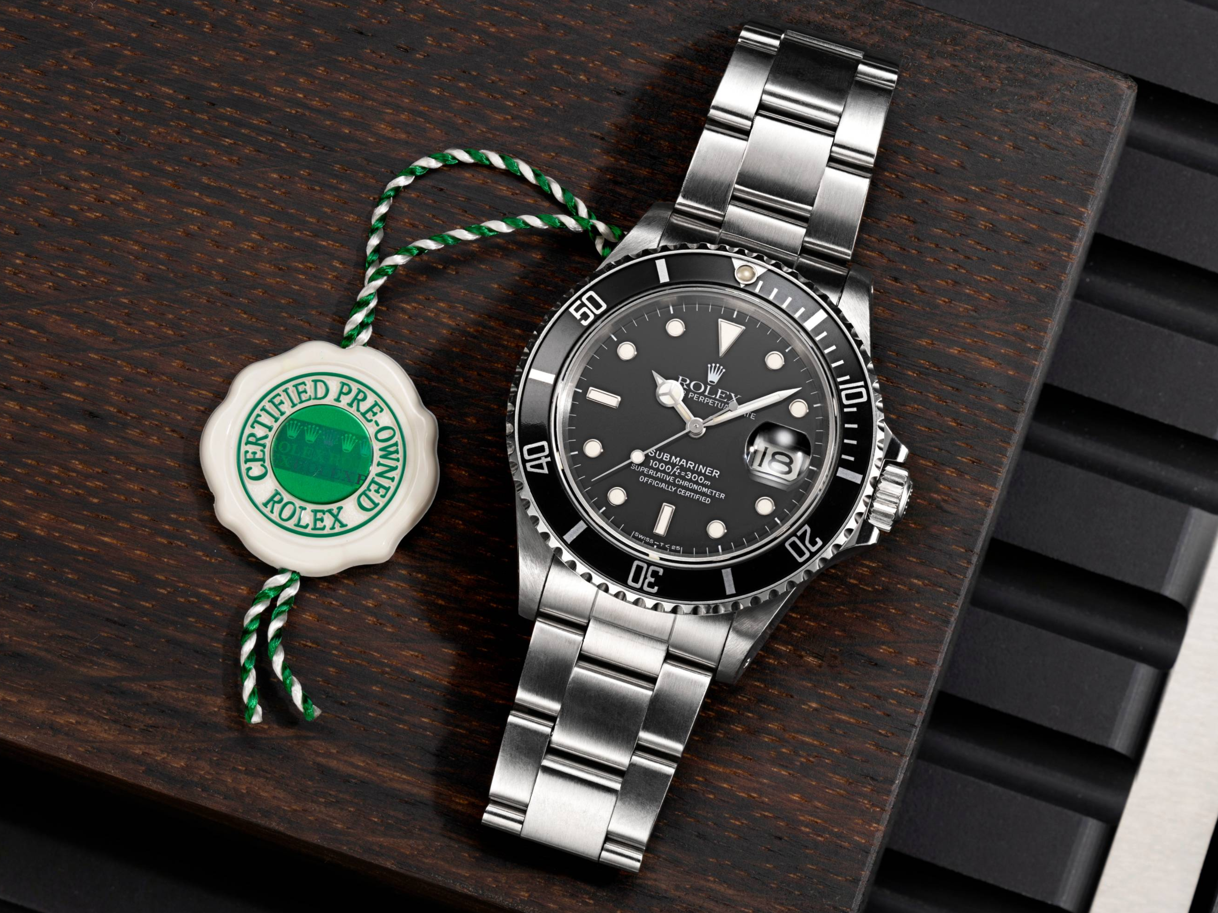 Une montre Rolex Submariner argentée avec cadran et lunette noirs, à côté d'une étiquette blanche et verte « Certifiée d'occasion ».