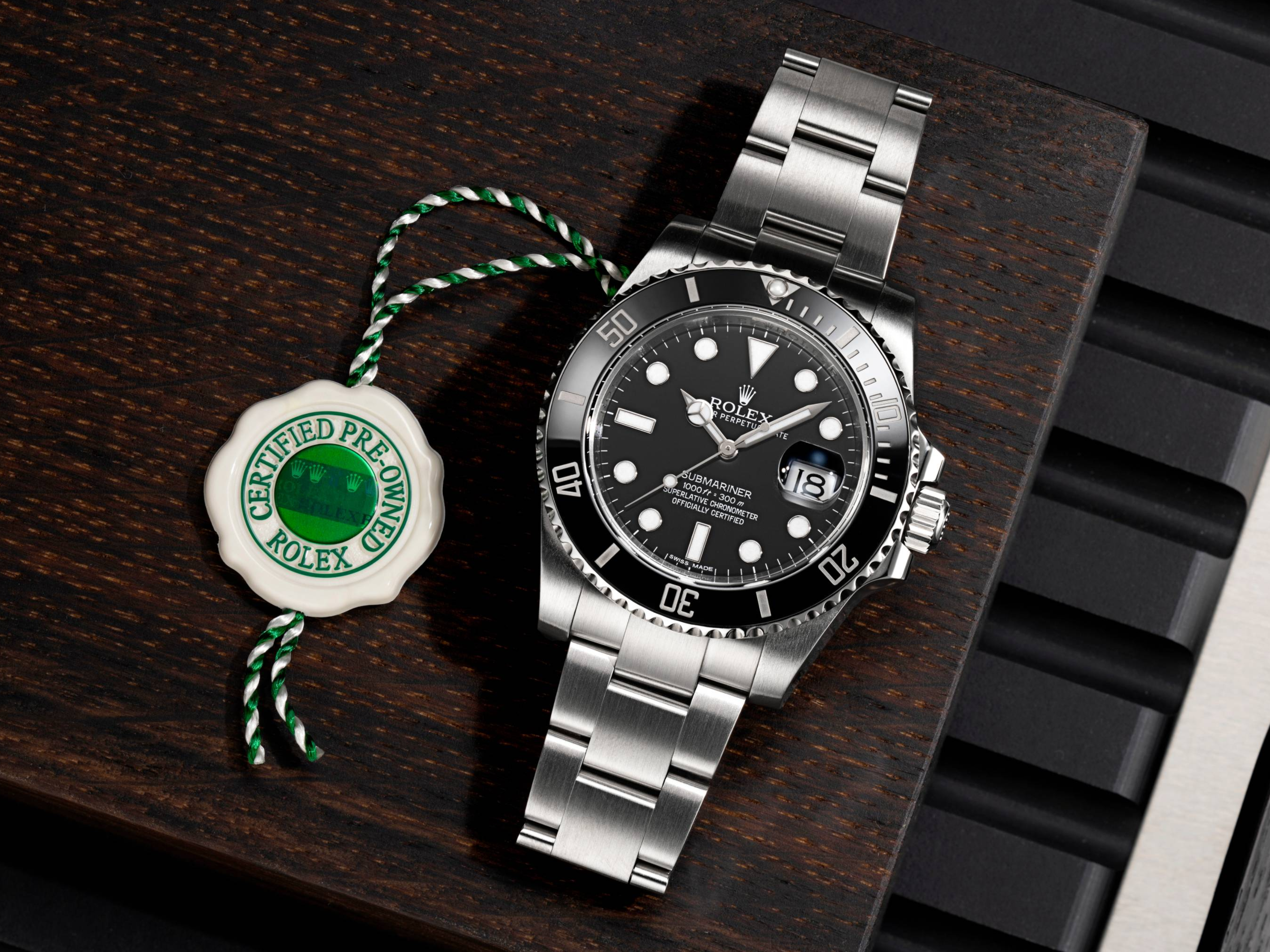 Une montre Rolex Submariner argentée avec cadran et lunette noirs, à côté d'un sceau blanc et vert « Rolex d'occasion certifiée », sur une surface en bois foncé.