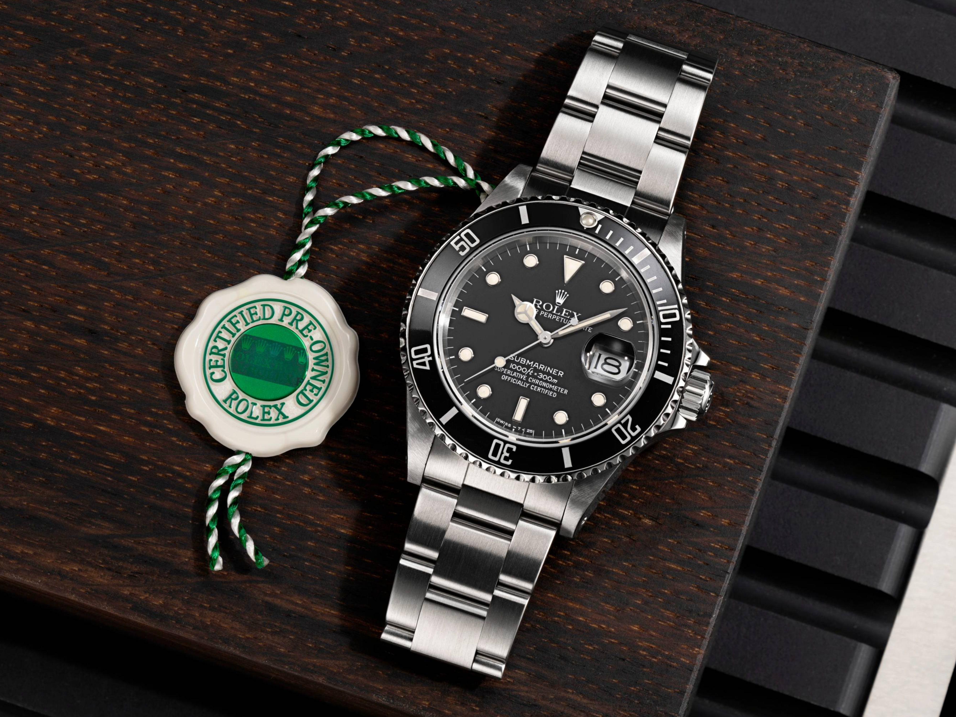 Une montre Rolex Submariner à cadran noir et bracelet en acier, à côté d'une étiquette « Certifiée d'occasion », sur une surface en bois foncé.