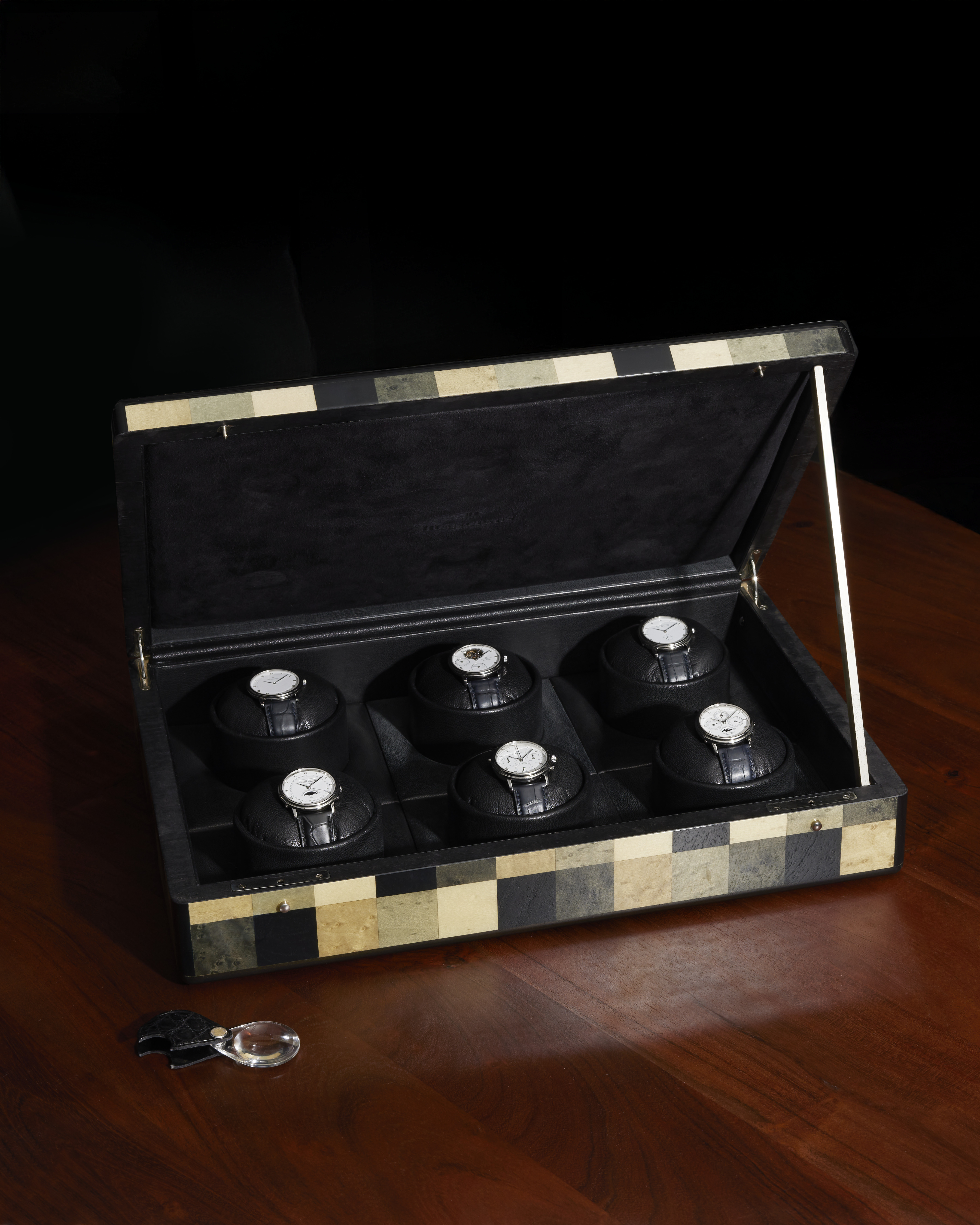 An open, black and white checkered watch box with six watches on display, next to a magnifying glass.