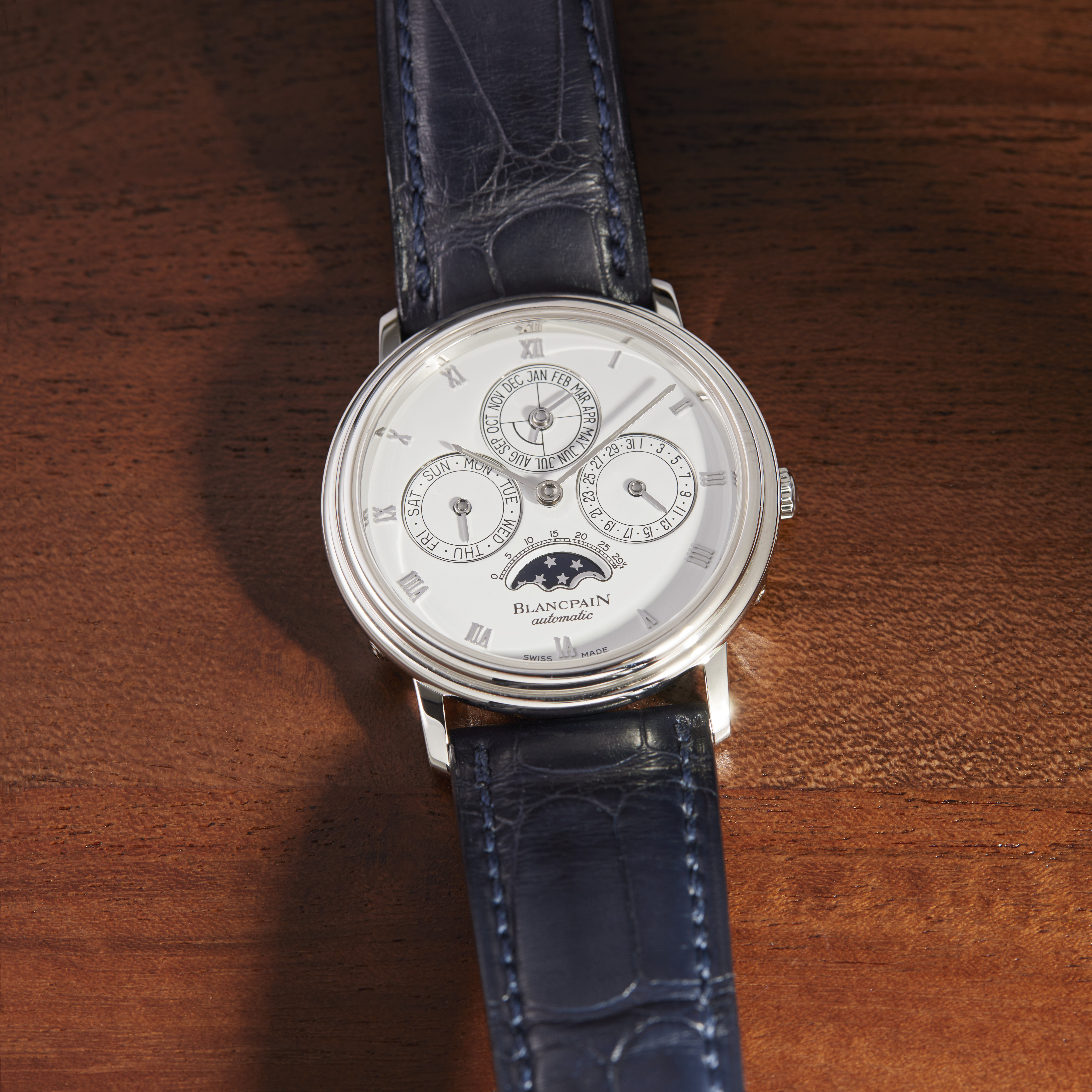 A silver Blancpain automatic watch with a white face, multiple subdials, and a dark blue alligator leather strap.