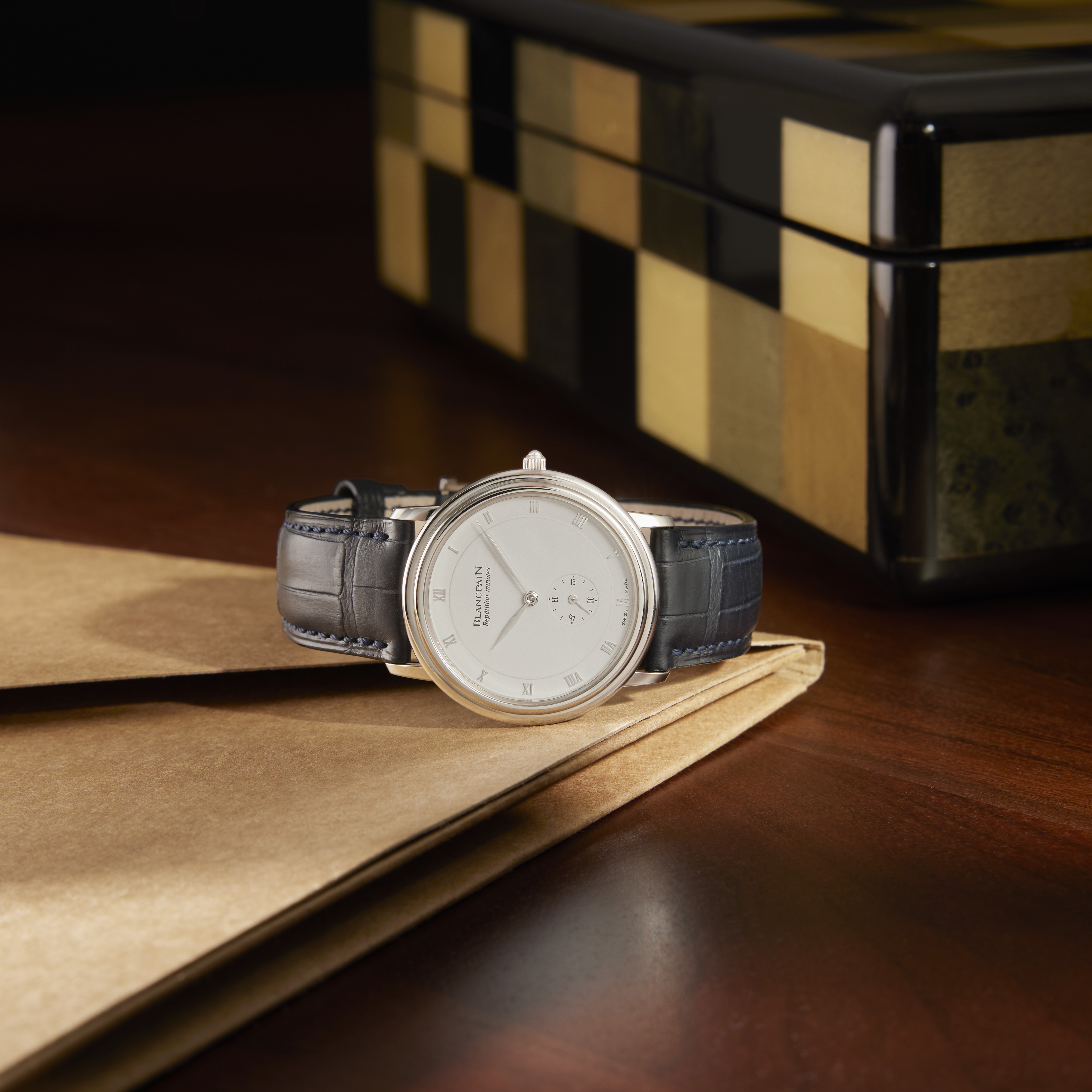A silver Blancpain watch with a white face and blue alligator strap rests on a brown folder, with a black and gold box in the background.