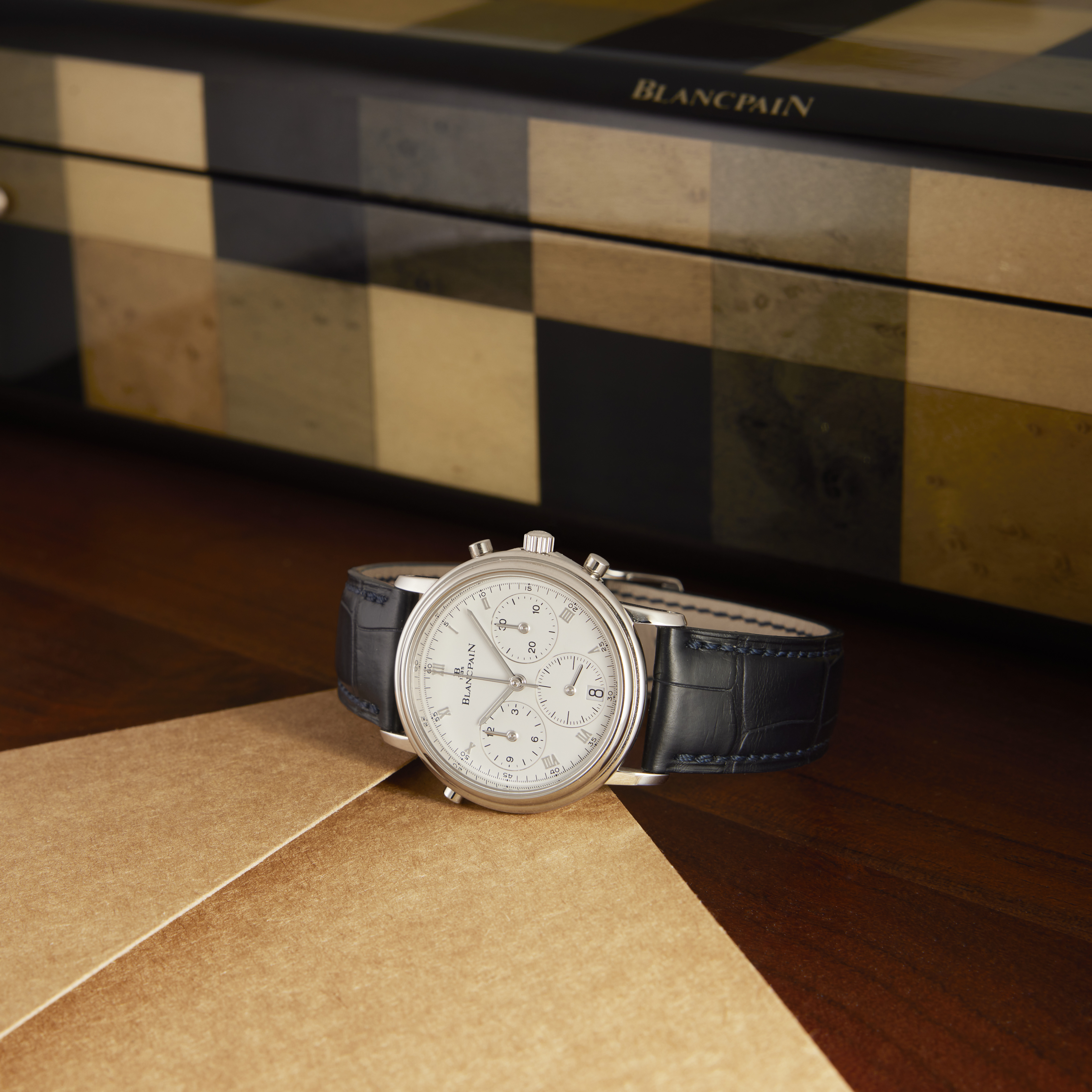 A silver Blancpain chronograph watch with a white dial and blue alligator strap on a wooden surface.