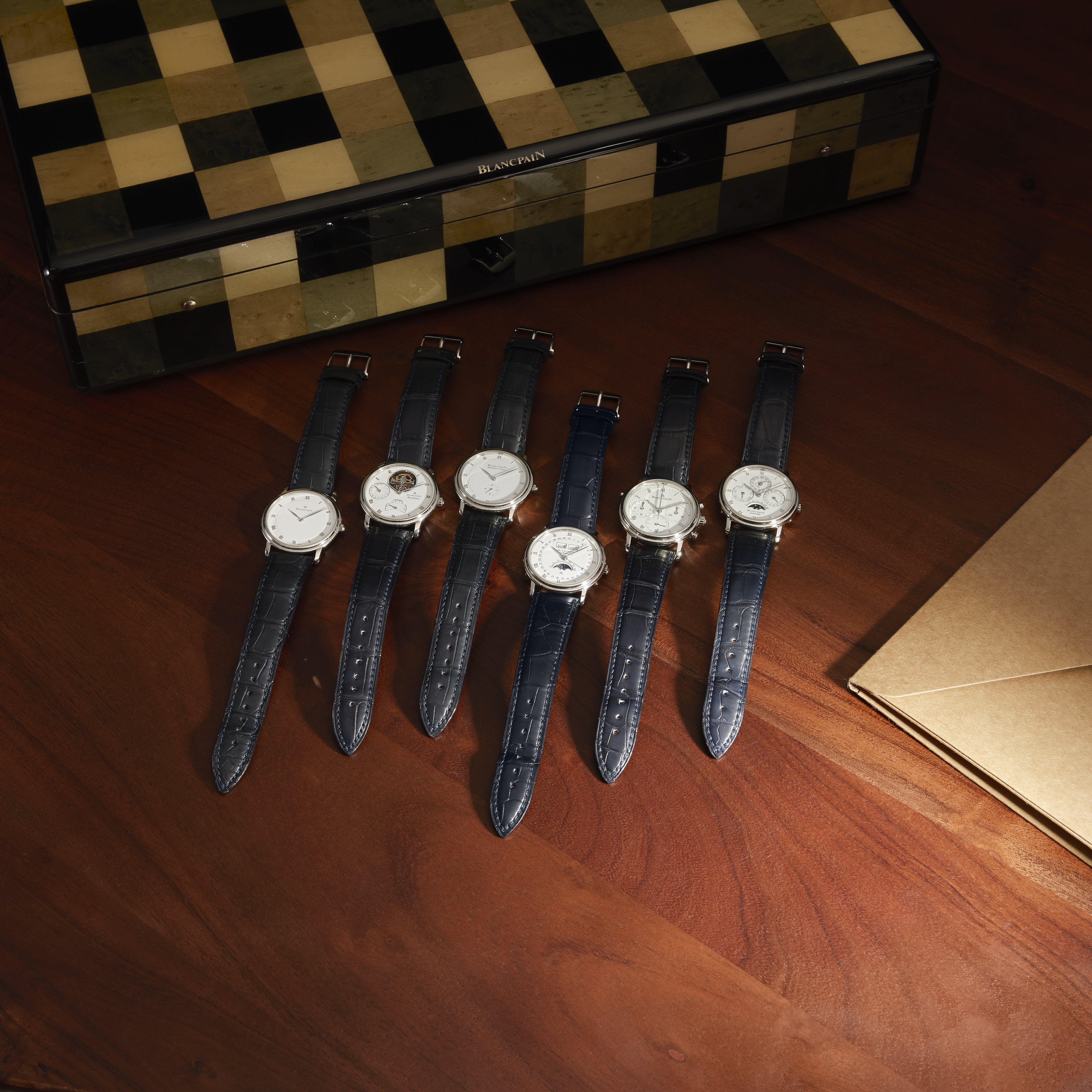 Six silver watches with dark blue leather bands arranged on a wooden table next to a checkered box and a brown envelope.