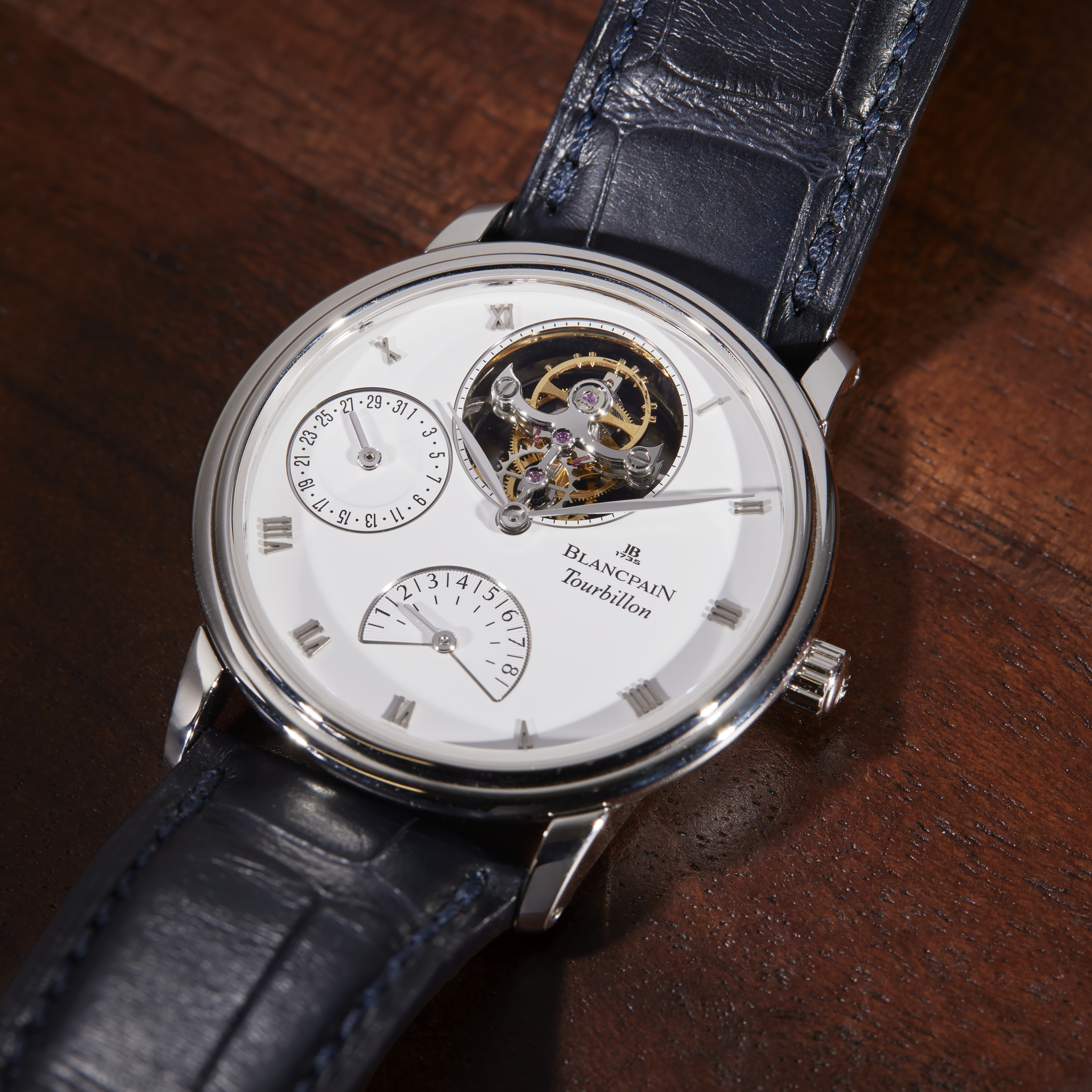 A silver Blancpain Tourbillon watch with a white face, exposed gears, and a dark blue leather strap on a wooden surface.