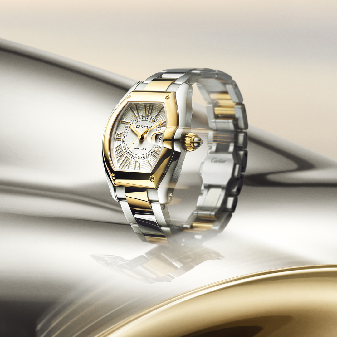 A gold and silver Cartier watch with a white face and Roman numerals, on a reflective metallic surface.