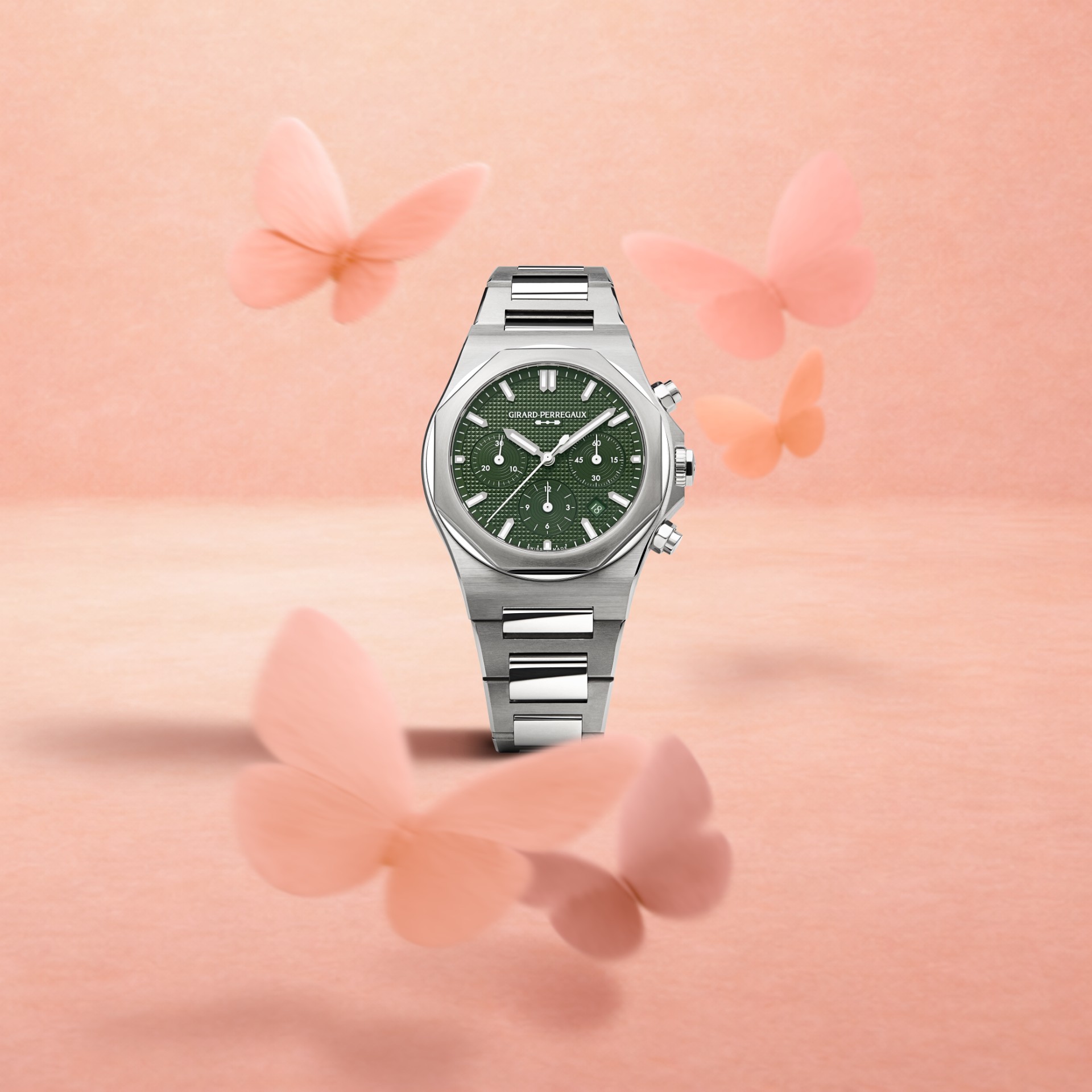 A silver watch with a green face and three subdials, surrounded by several blurred pink butterflies.