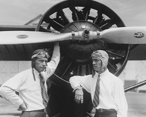 Two male pilots in flight helmets and goggles stand beside a large propeller plane.