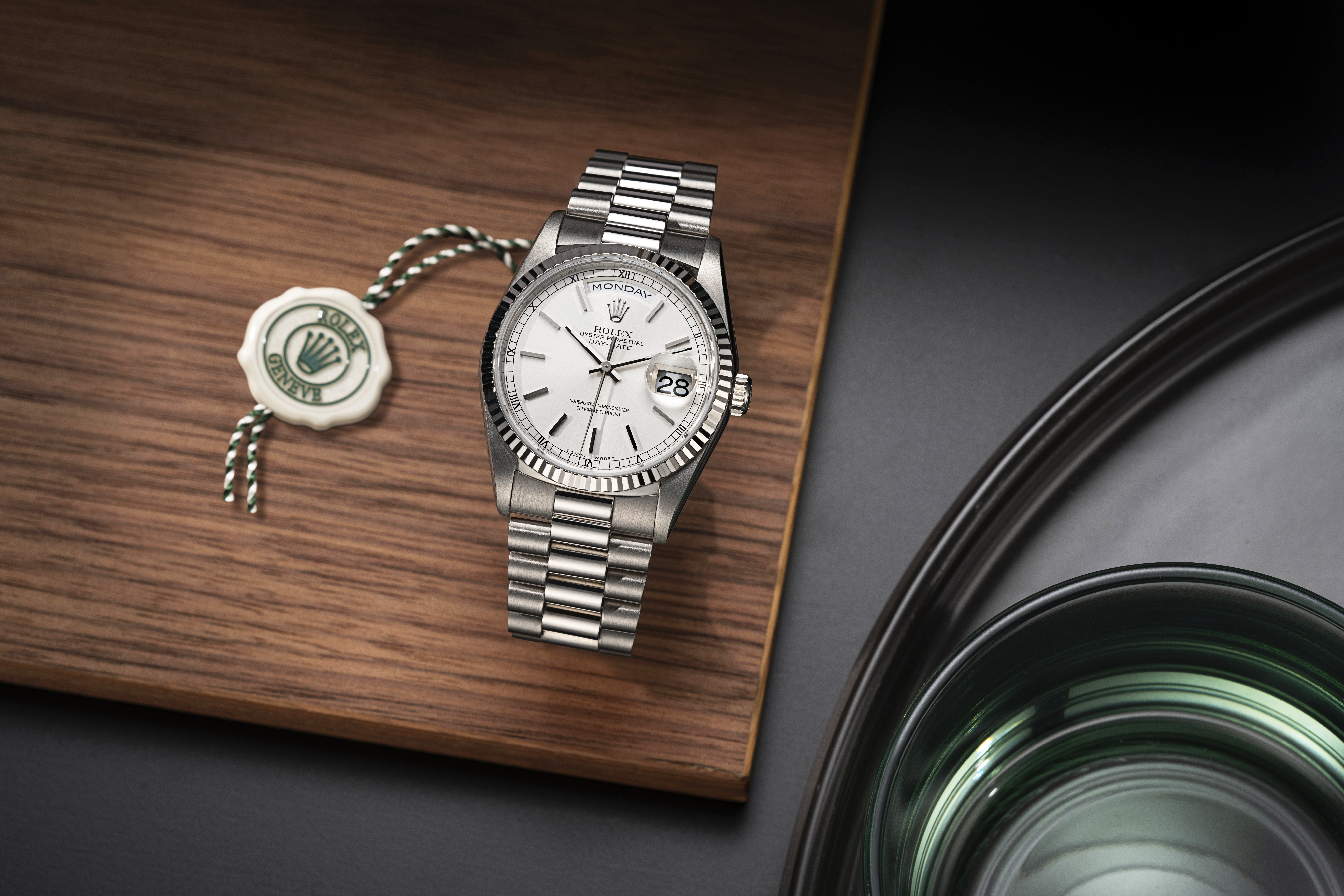 Une montre Rolex Day-Date argentée, à cadran blanc et sceau de cire verte, posée sur une planche en bois.