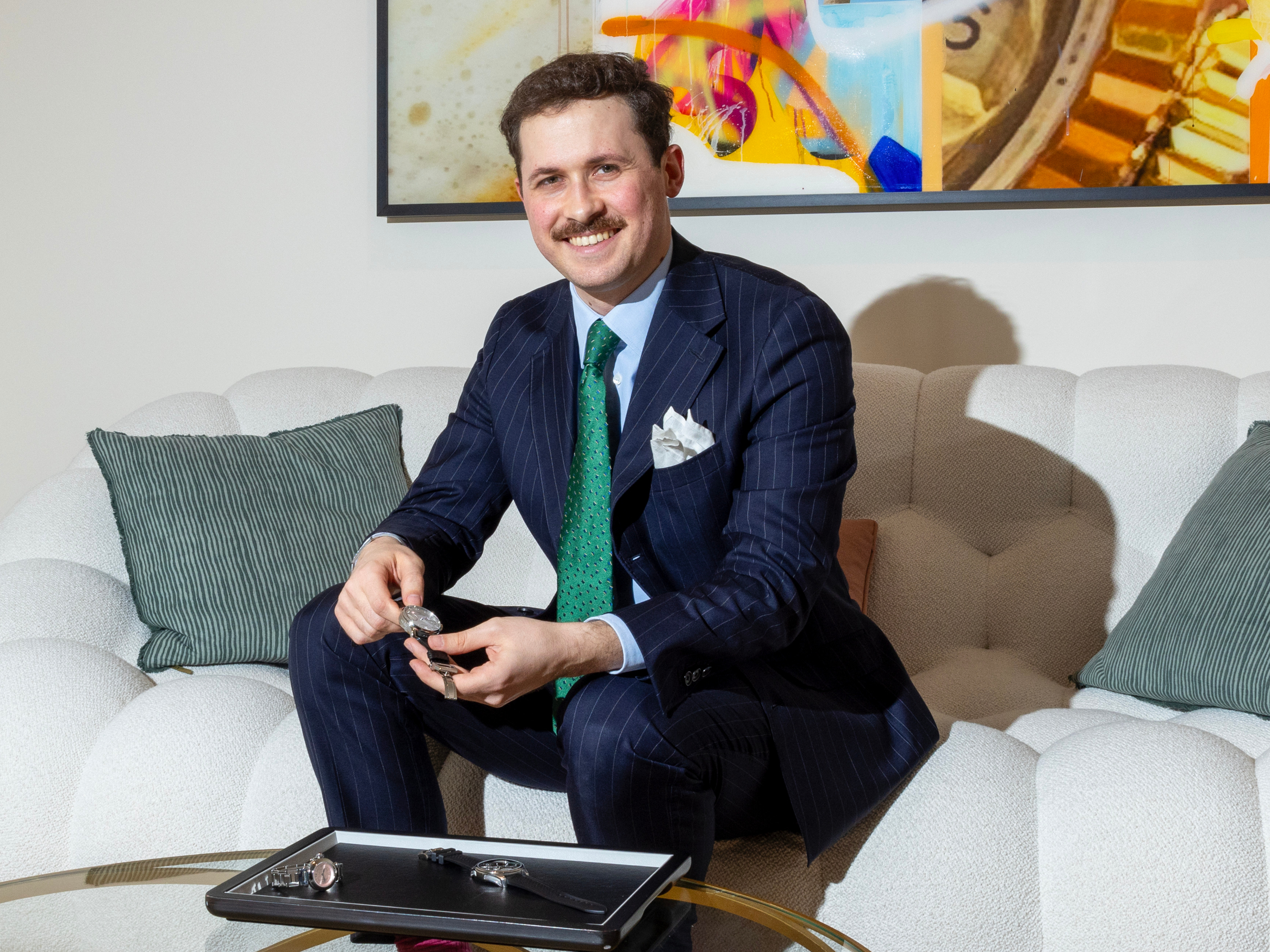 A man in a pinstripe suit and green tie sits on a white sofa, holding a watch and smiling.