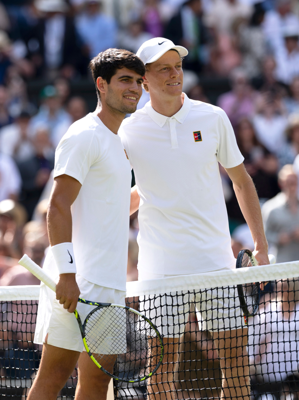 Two male tennis players in white outfits stand on a court, smiling.