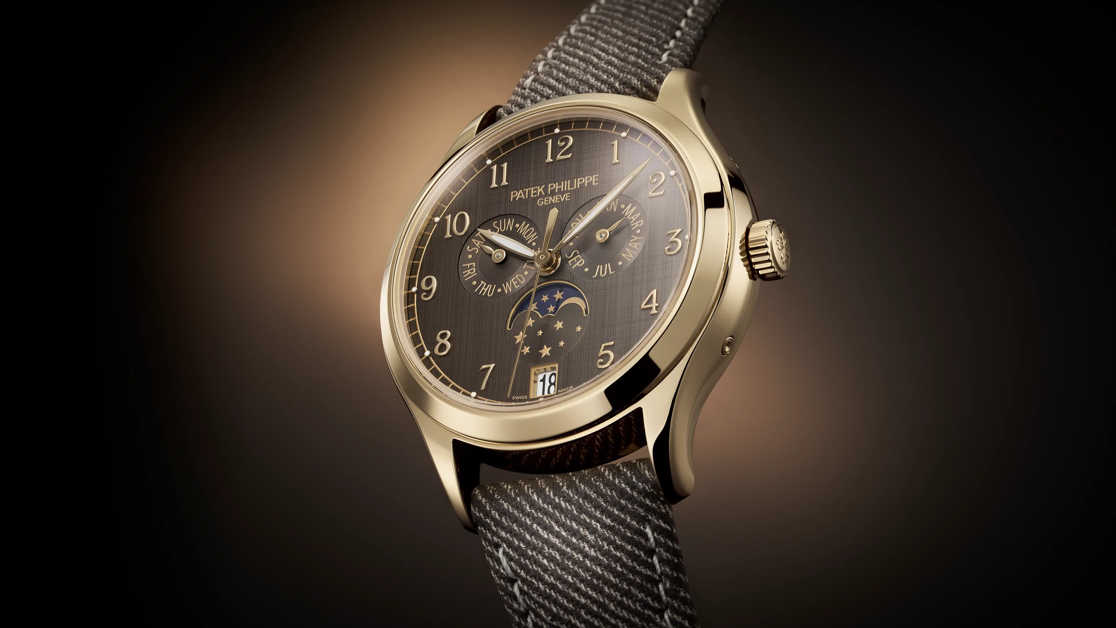 A gold Patek Philippe watch with a dark gray face, moon phase, and calendar complications on a striped fabric strap.