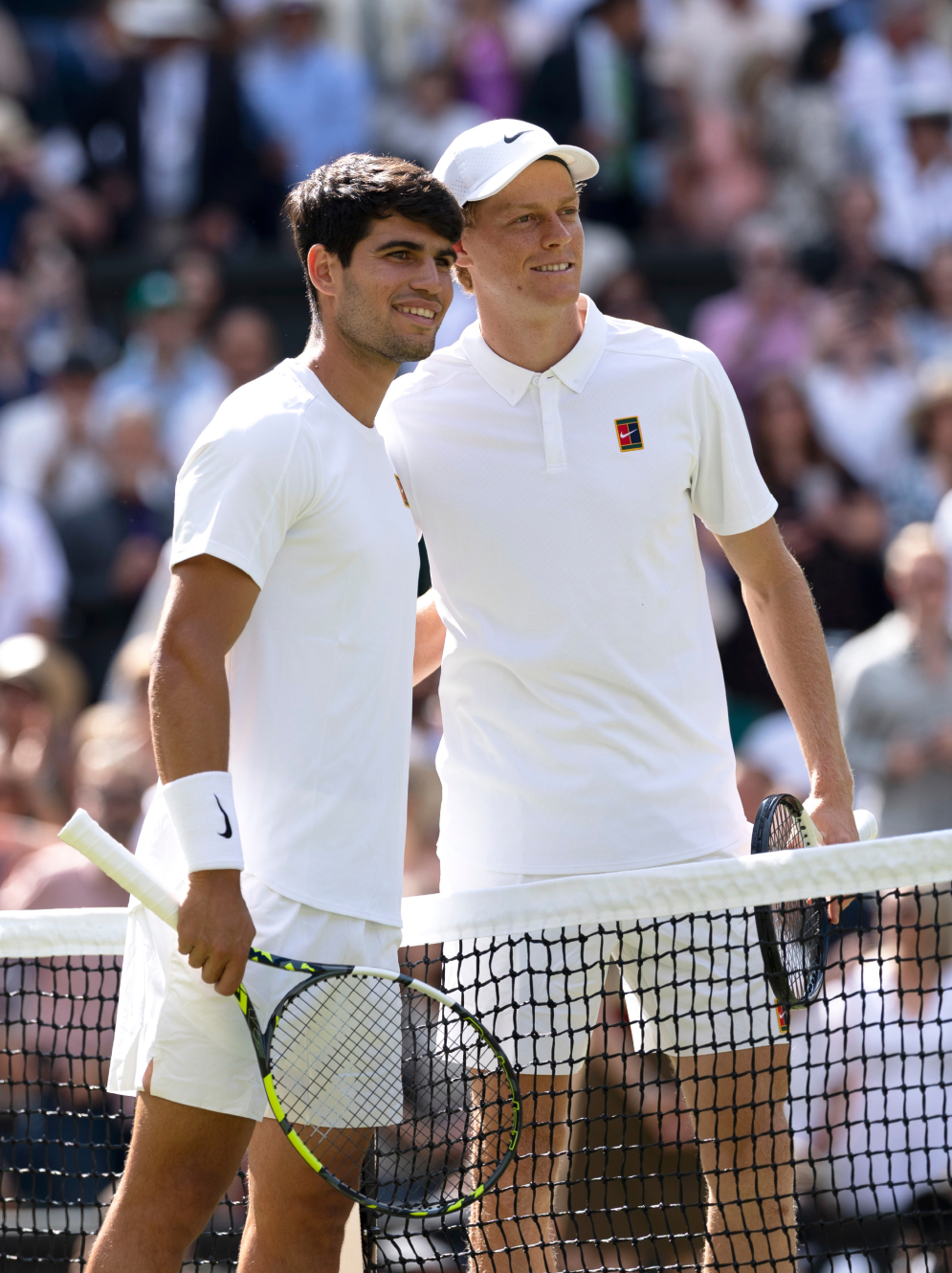 Zwei männliche Tennisspieler in weißen Outfits, die Schläger haltend, stehen lächelnd auf einem Platz, zwischen ihnen befindet sich ein Netz.