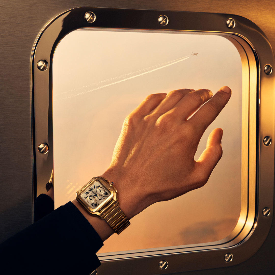 A gold watch on a person's wrist, with their hand reaching towards an airplane window showing a plane in the sky.