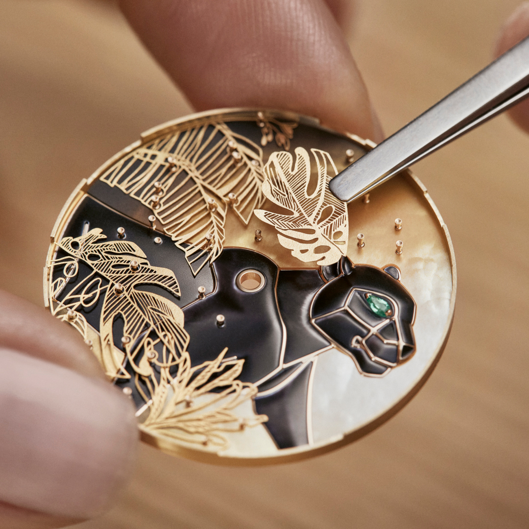 A close-up of a gold and black medallion with a panther and tropical leaves, being worked on by tweezers.