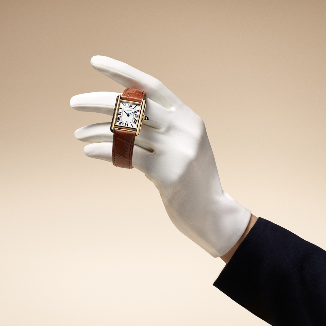 A gloved hand holds a gold rectangular watch with a white face and brown leather strap.