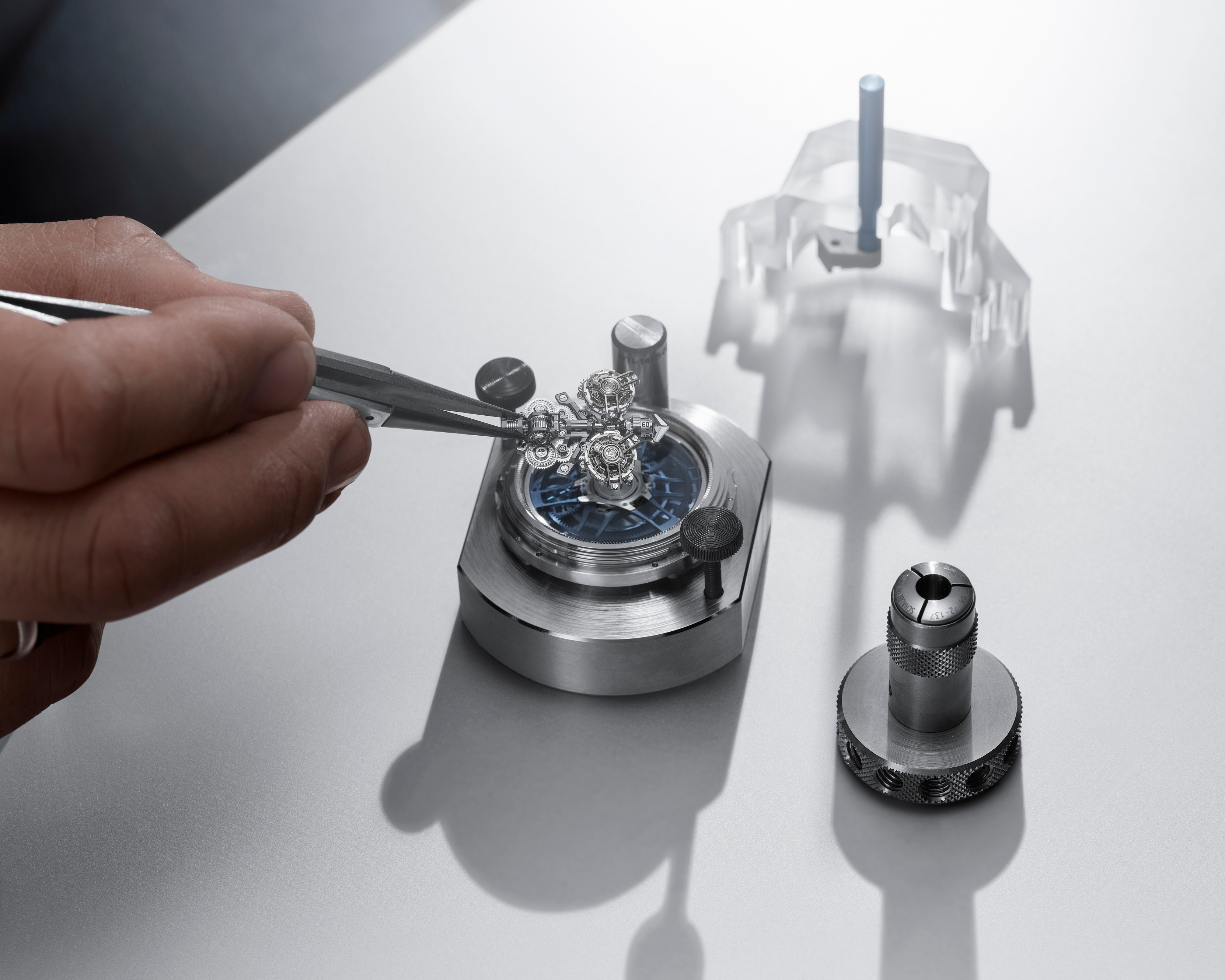 A person's hand uses tweezers to place a tiny watch mechanism onto a metal base, with other tools nearby.