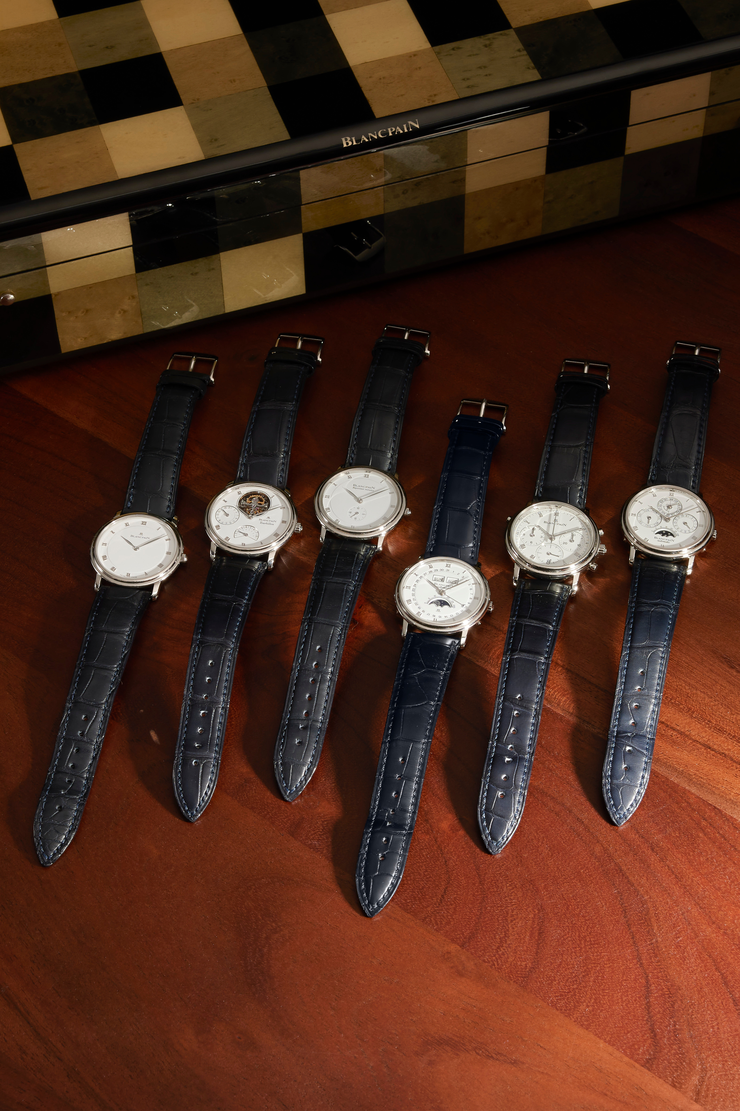 Sechs Blancpain-Uhren mit weißen Zifferblättern und dunkelblauen Lederarmbändern sind auf einer Holzfläche vor einer schwarz-braun karierten Schachtel angeordnet.